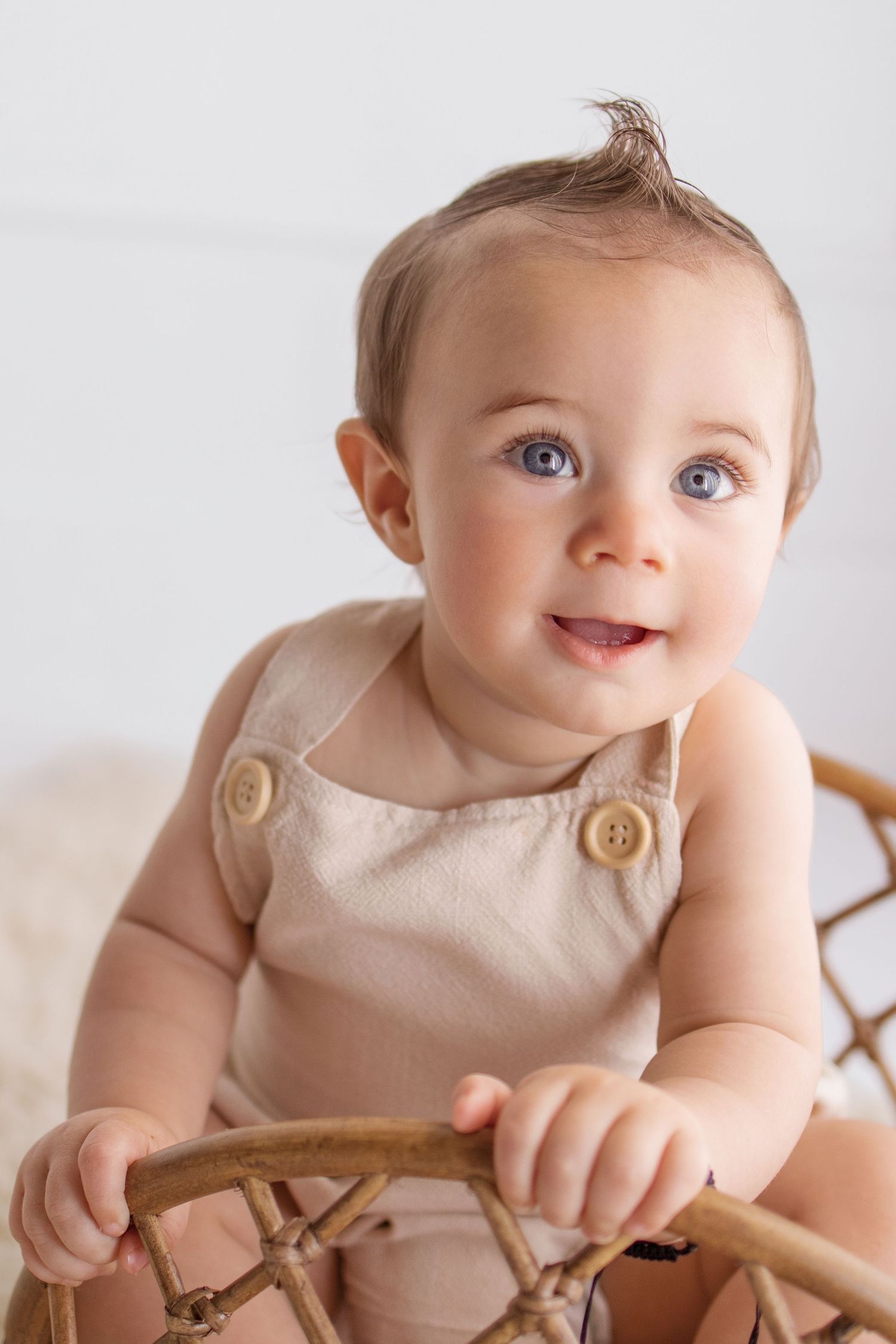 Smiling baby with blue eyes wearing a beige linen romper sitting in a rattan chair against a white background.