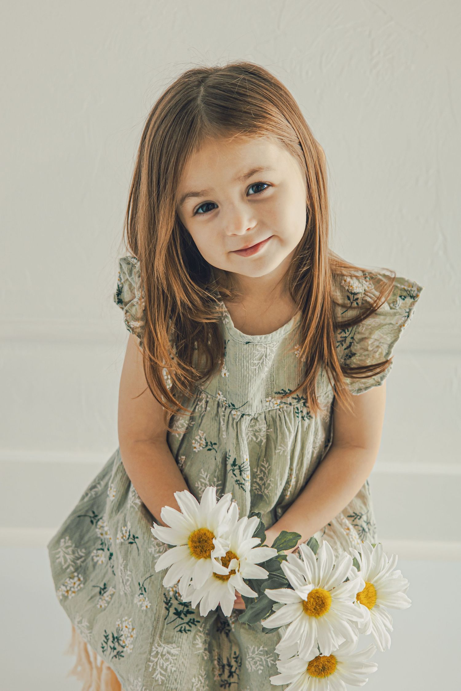 Young girl in floral dress holding white daisies, smiling softly at camera in bright studio setting.