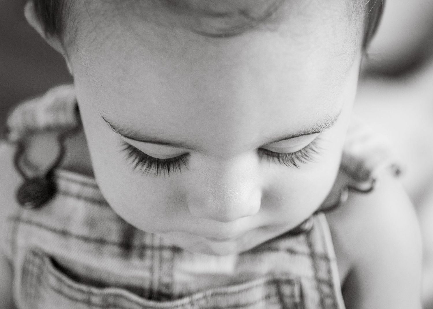 Close-up black and white portrait of a baby with long eyelashes looking down, wearing overalls.