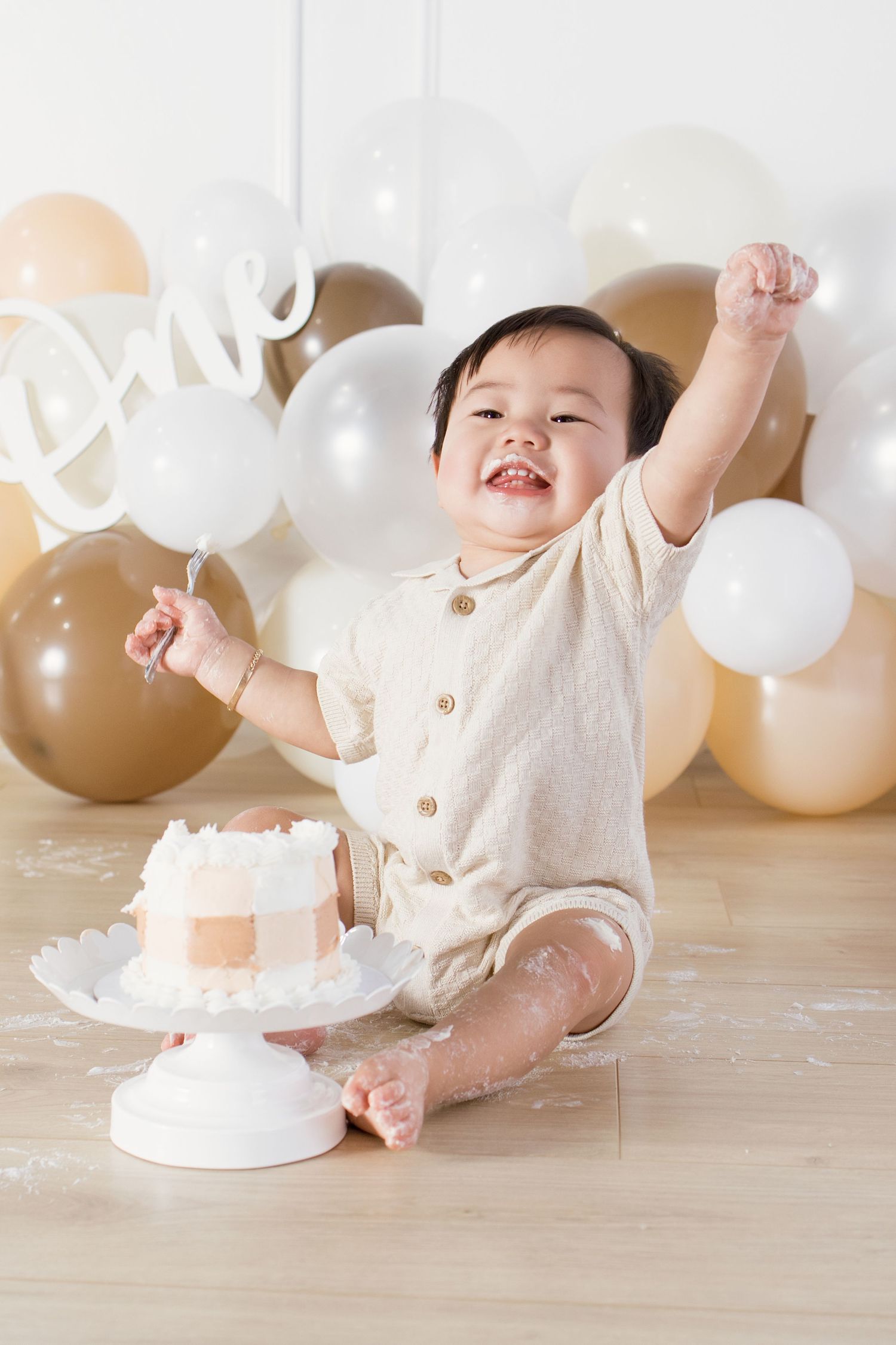 A joyful baby celebrates their first birthday sitting on the floor with a smash cake, surrounded by gold and white balloons.