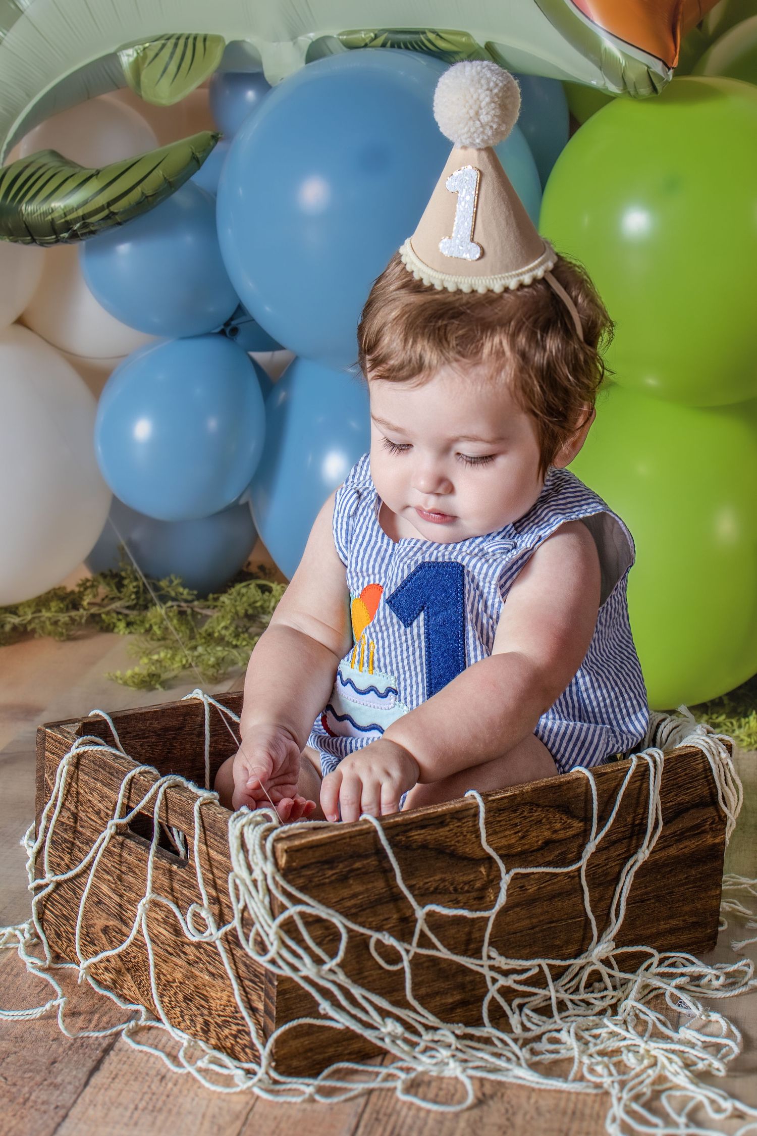 Baby boy in birthday hat sits in wooden crate surrounded by blue and green balloons at first birthday party.