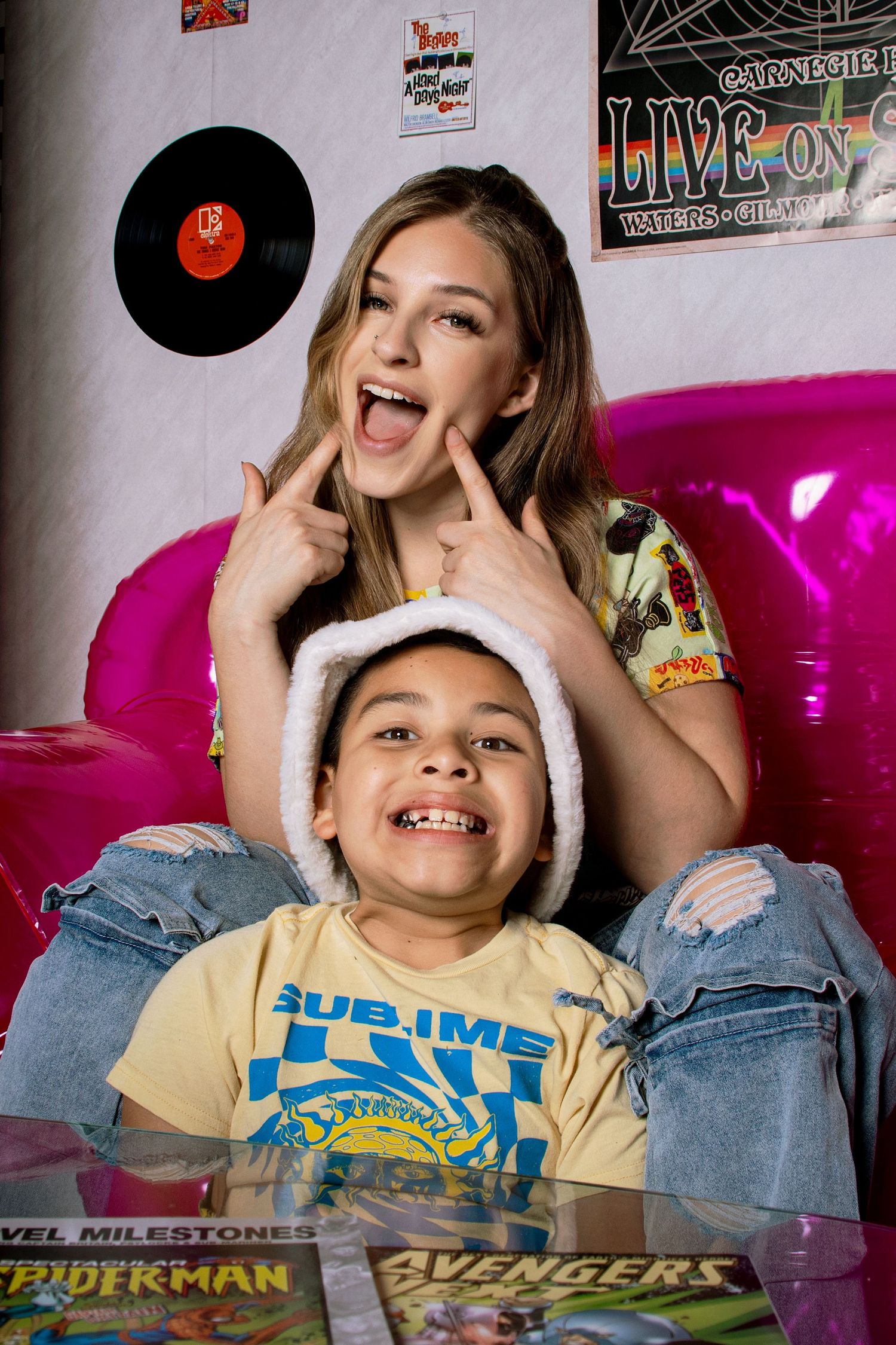 A laughing woman points to her cheeks while posing with a smiling boy in a Santa hat amid comic books.
