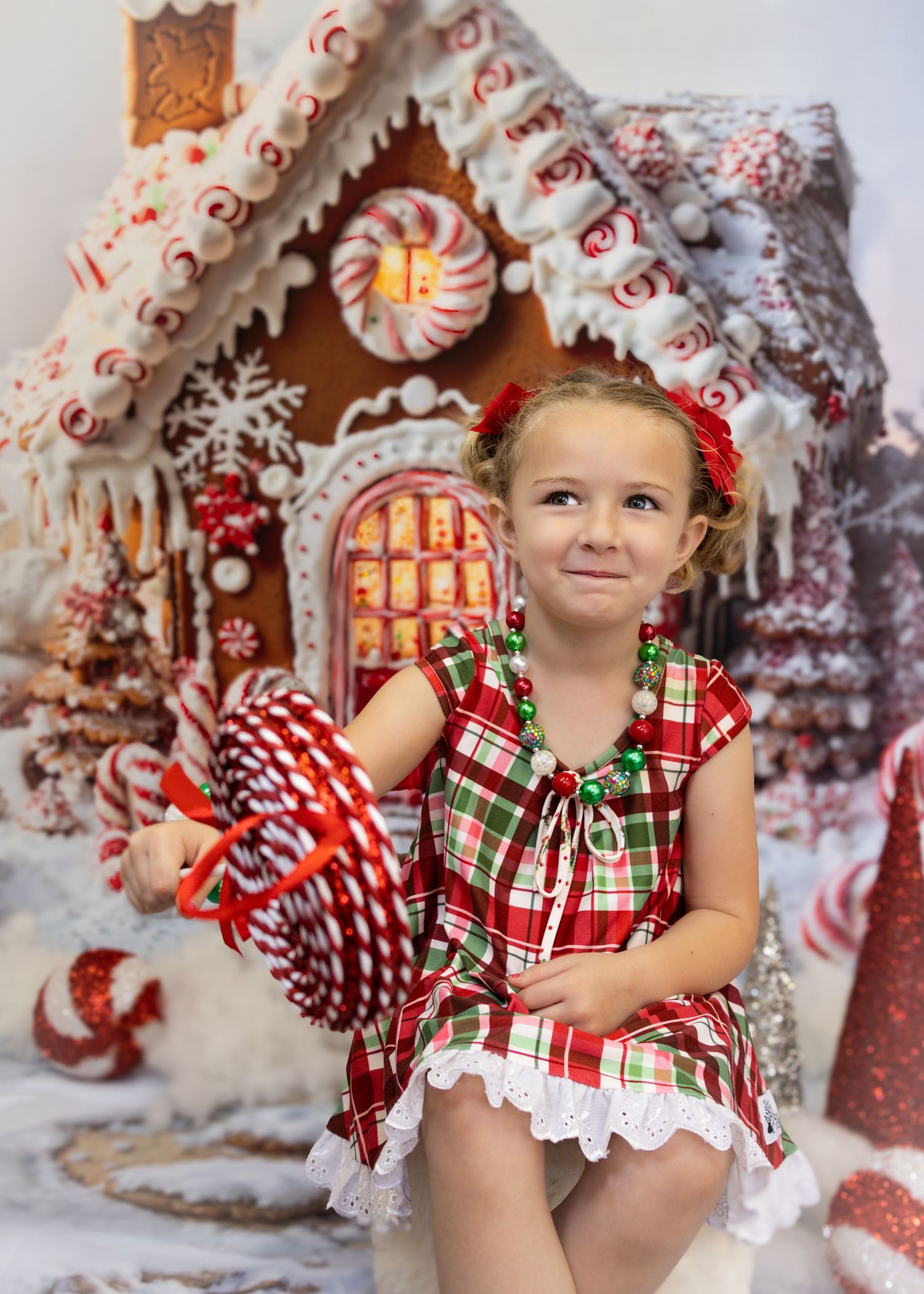 Young girl in plaid dress holds candy cane beside decorated gingerbread house with festive Christmas decor.