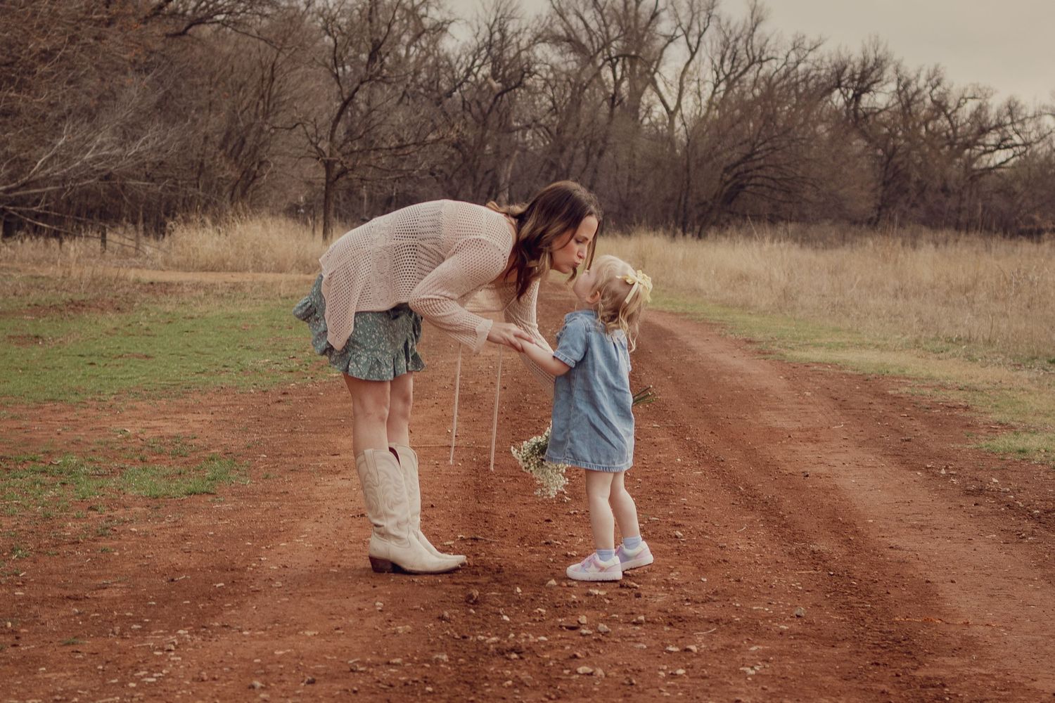 A mother bends down to interact with her young daughter on a rustic dirt road surrounded by bare trees.