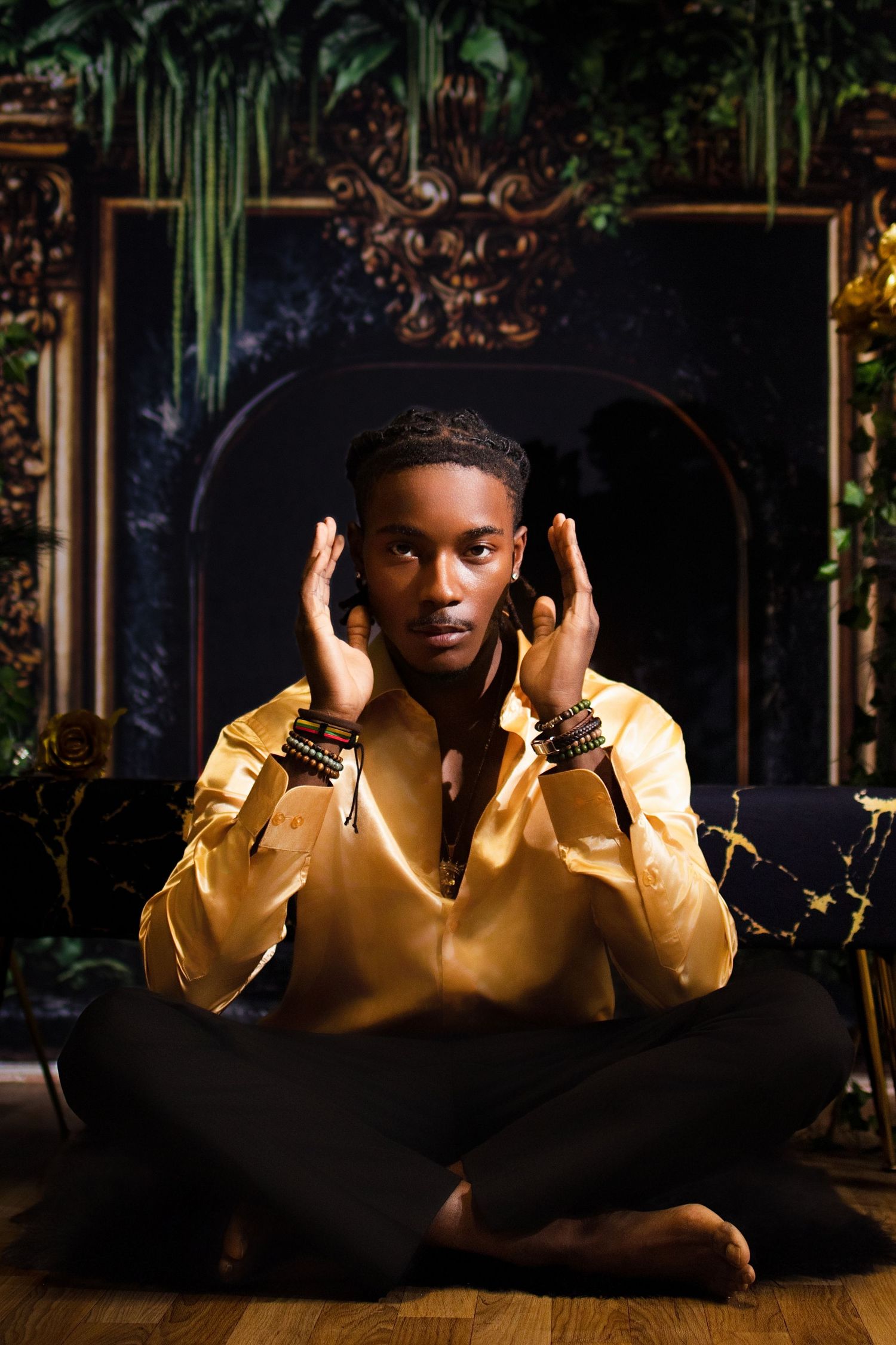 Young Black man in gold satin shirt sitting cross-legged on floor with hands raised in elegant pose.