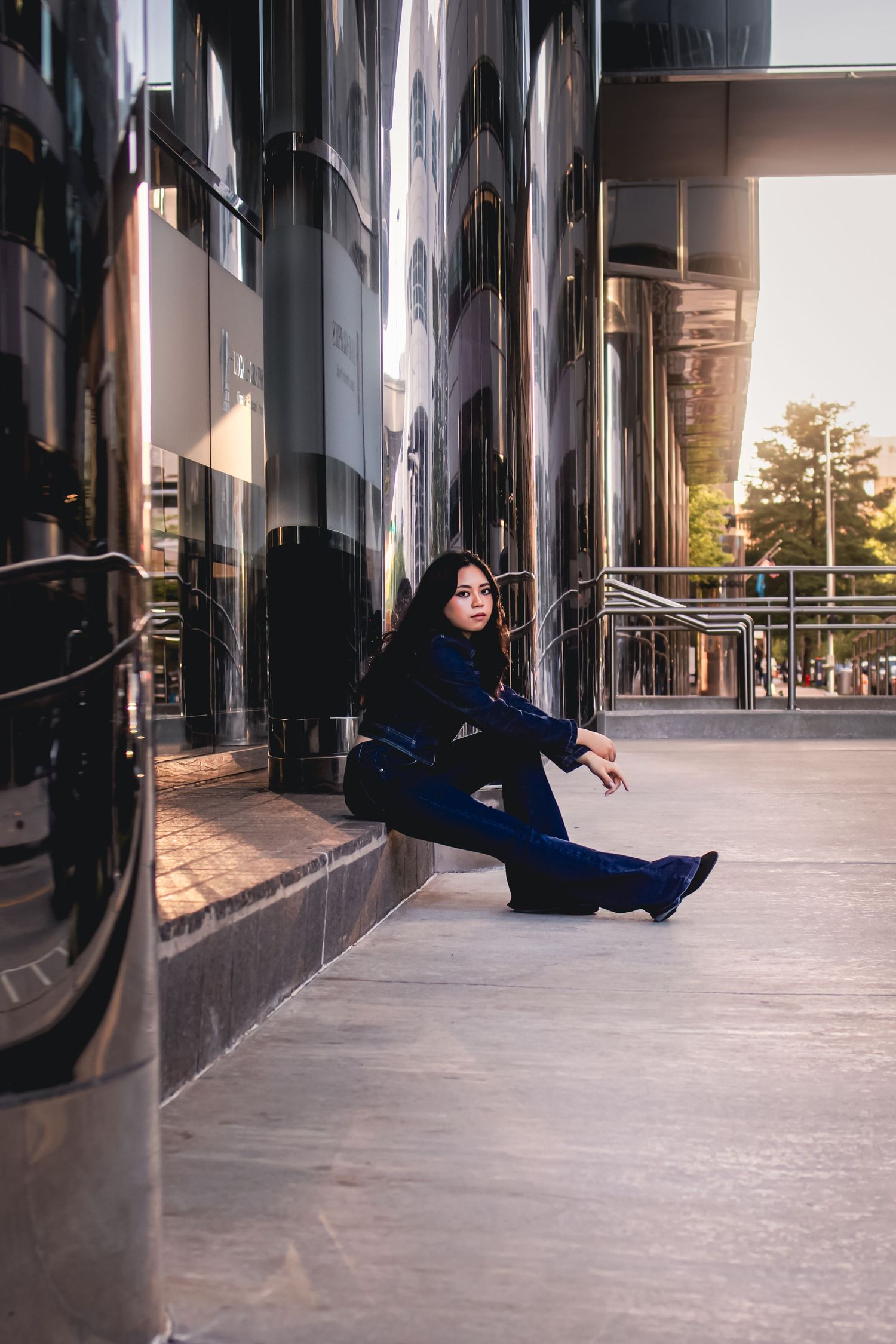 Young woman in blue outfit sits gracefully against reflective black building exterior in urban setting at golden hour.