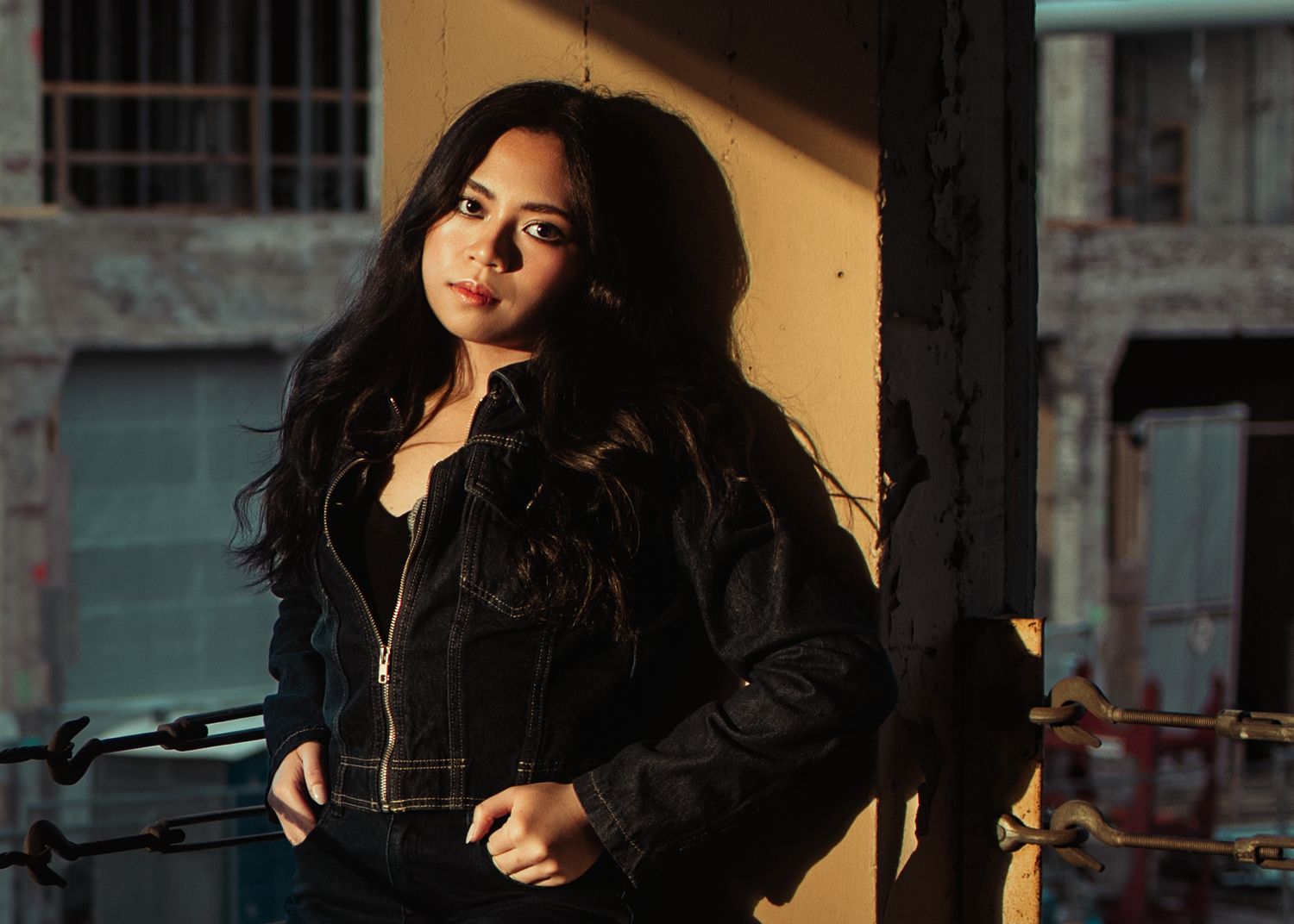 Young woman in black leather jacket posing confidently against urban building in dramatic natural lighting.