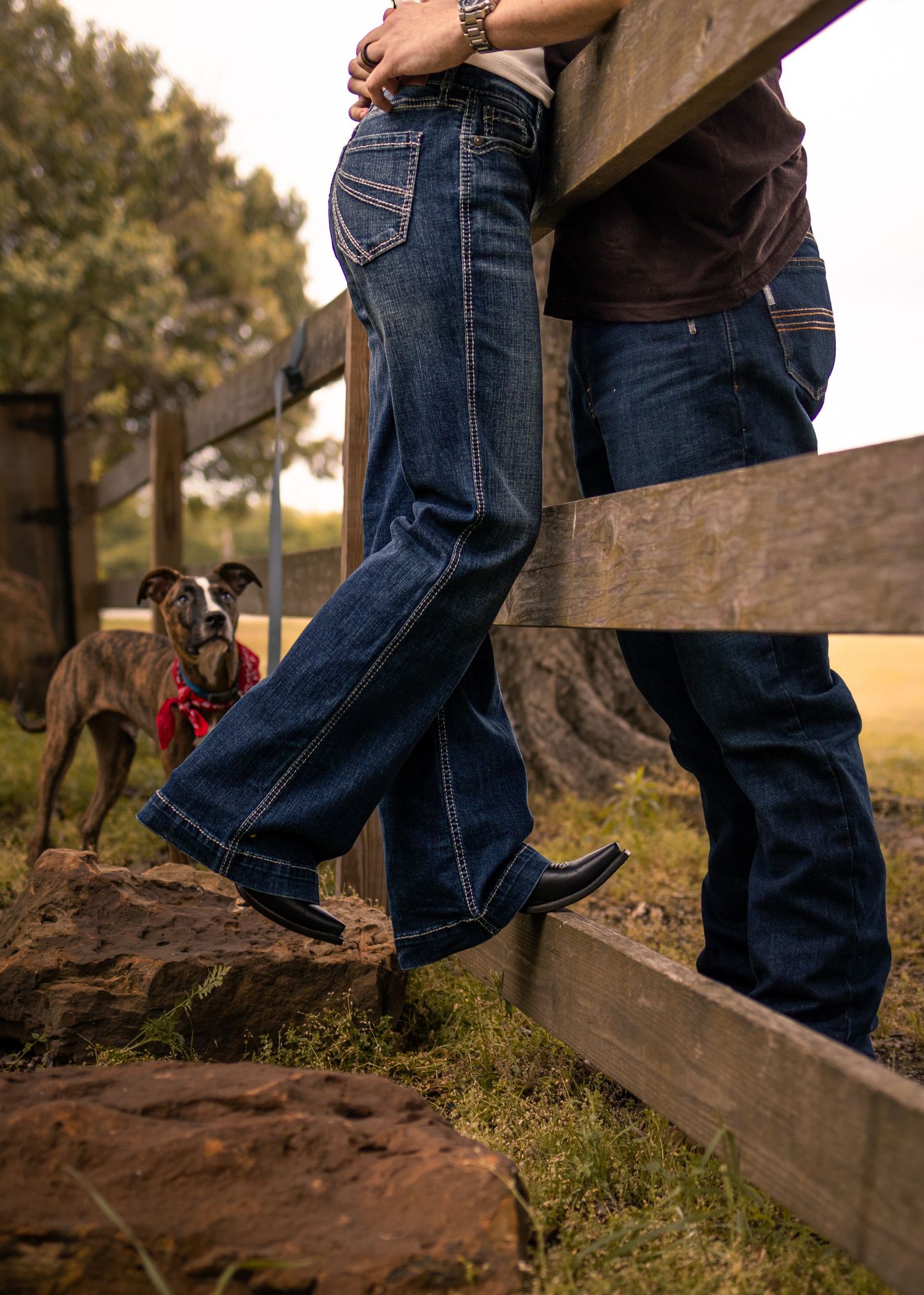 Couple in dark wash jeans and cowboy boots lean against a wooden fence with a greyhound dog nearby.