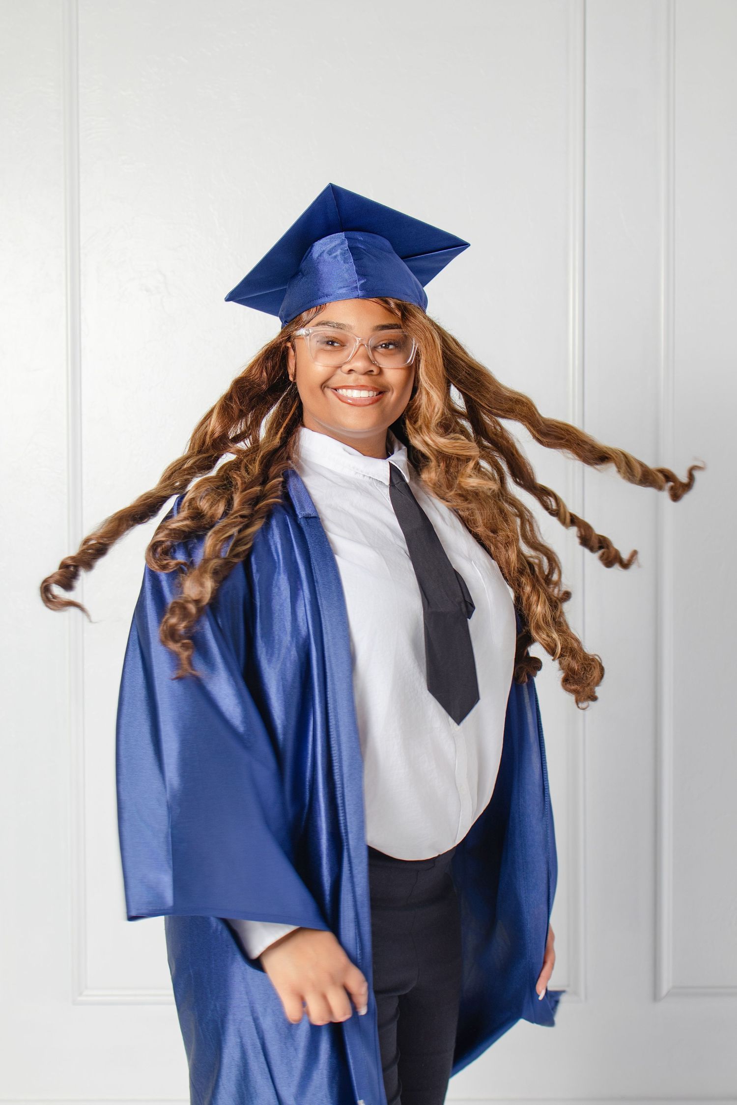 Joyful graduate in blue cap and gown with long curly hair flying, wearing glasses.