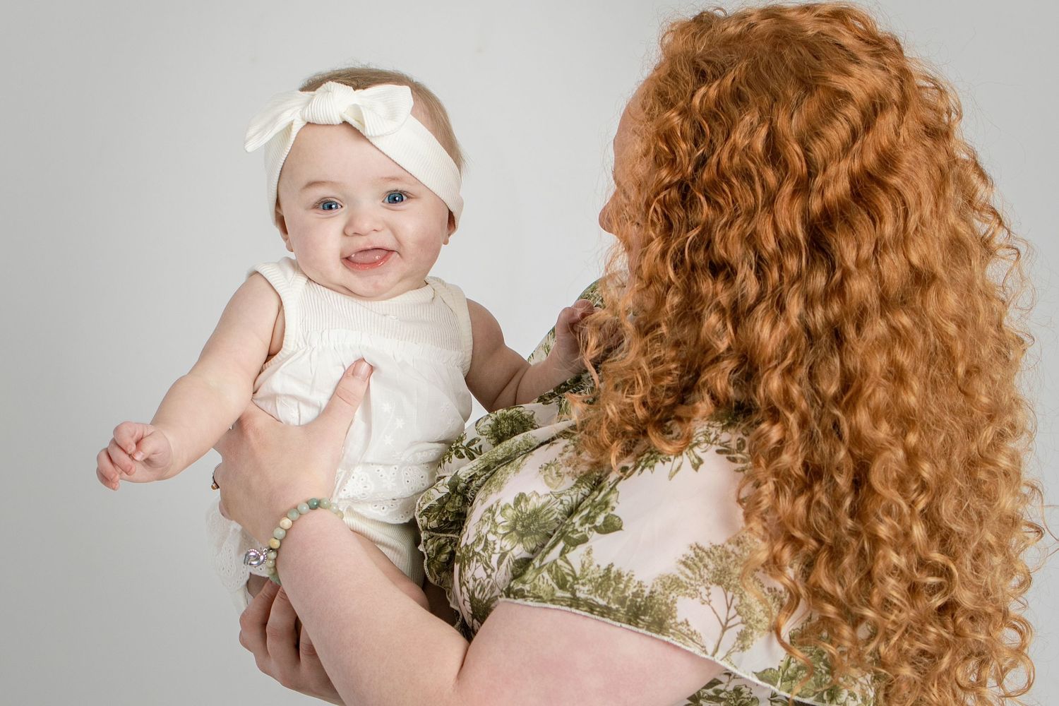 Smiling baby in white outfit held by redhead curly-haired woman in floral dress against gray background.