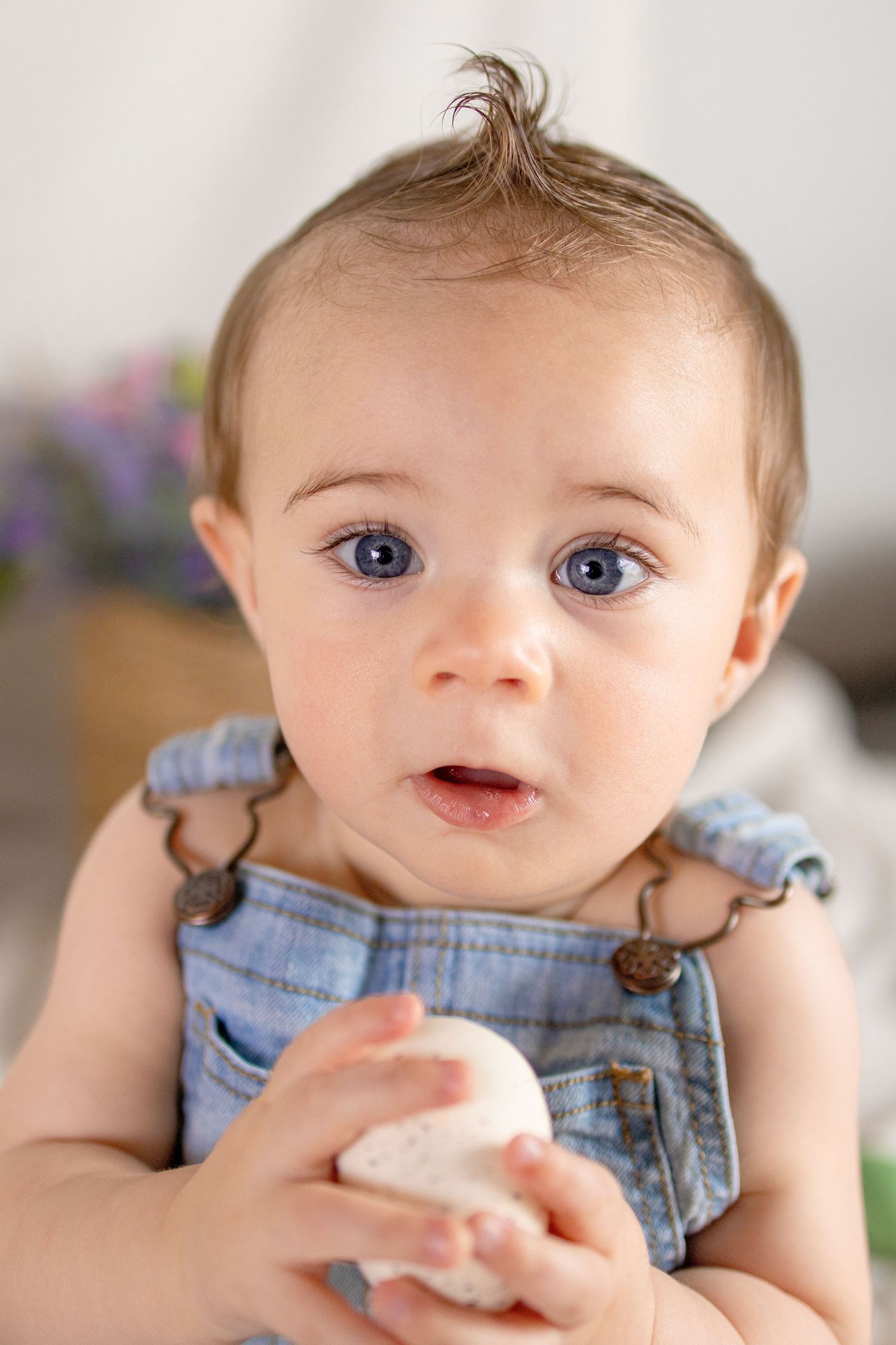 Adorable blue-eyed baby in denim overalls holding a white Easter egg.