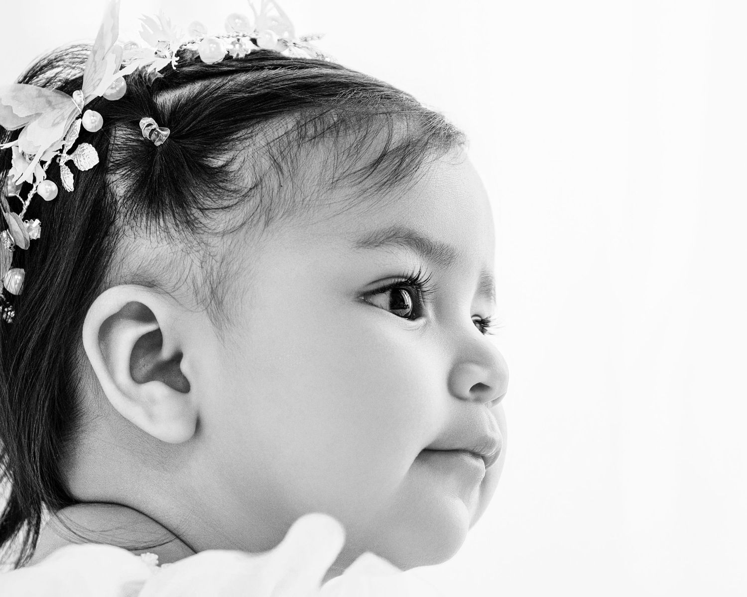 Black and white portrait of a young child wearing a floral headpiece, looking sideways with a gentle smile.