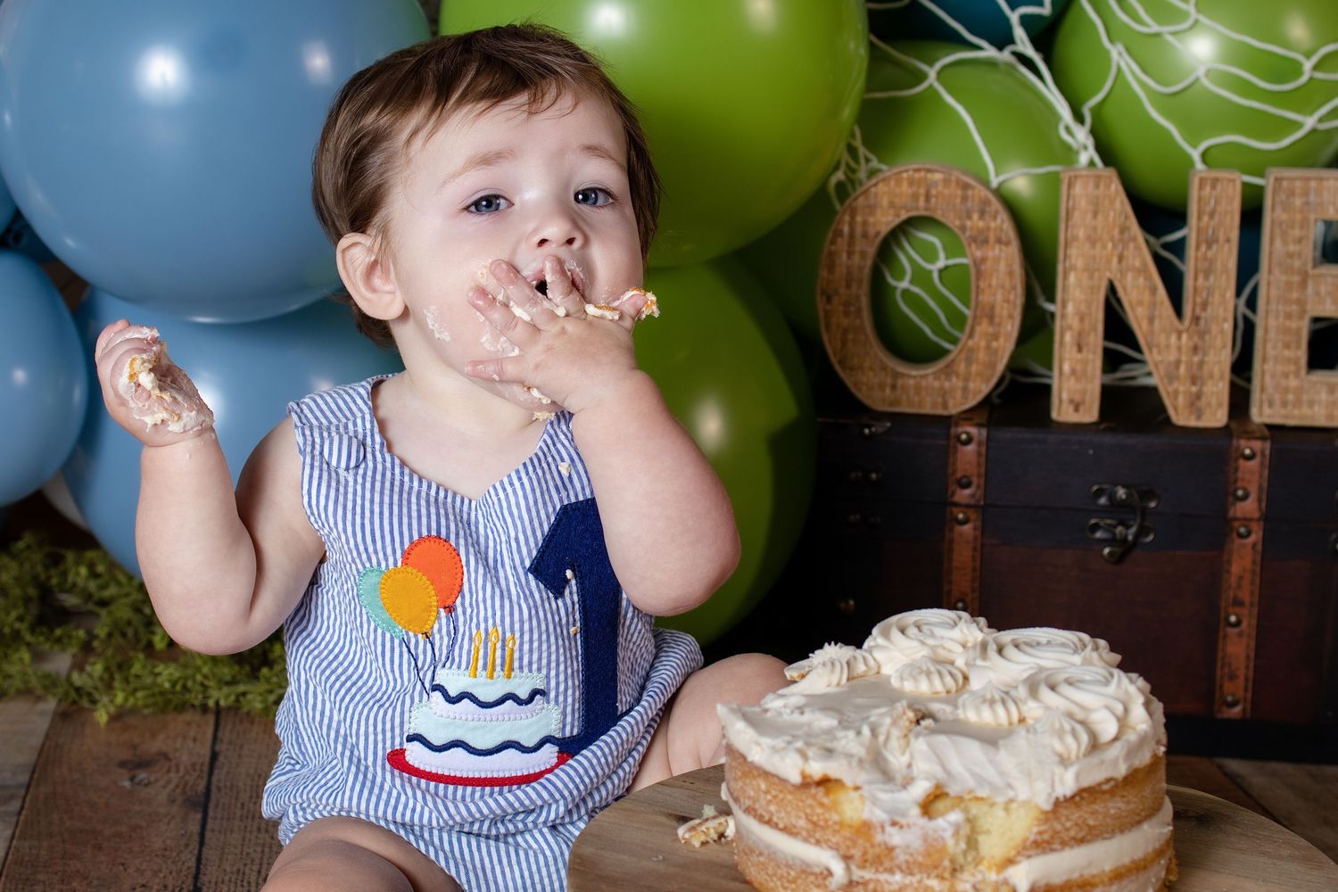 Baby boy smashes first birthday cake surrounded by blue and green balloons and ONE sign decor.