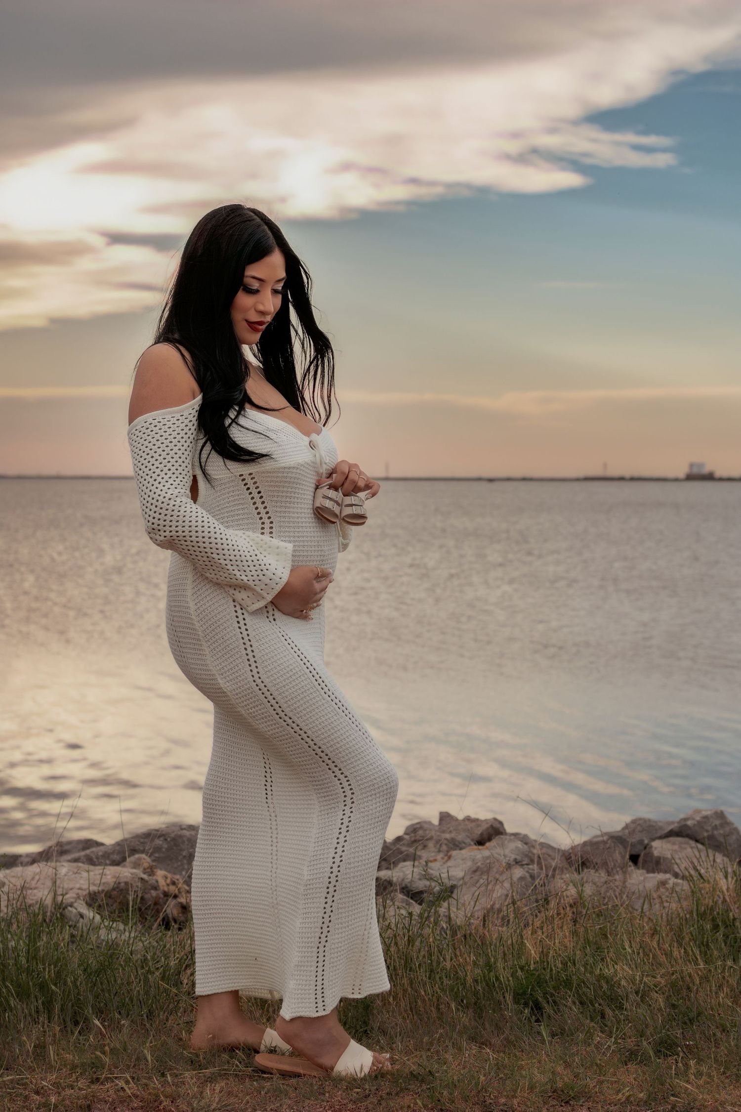 Pregnant woman in white knit dress holding baby shoes by the water at sunset.