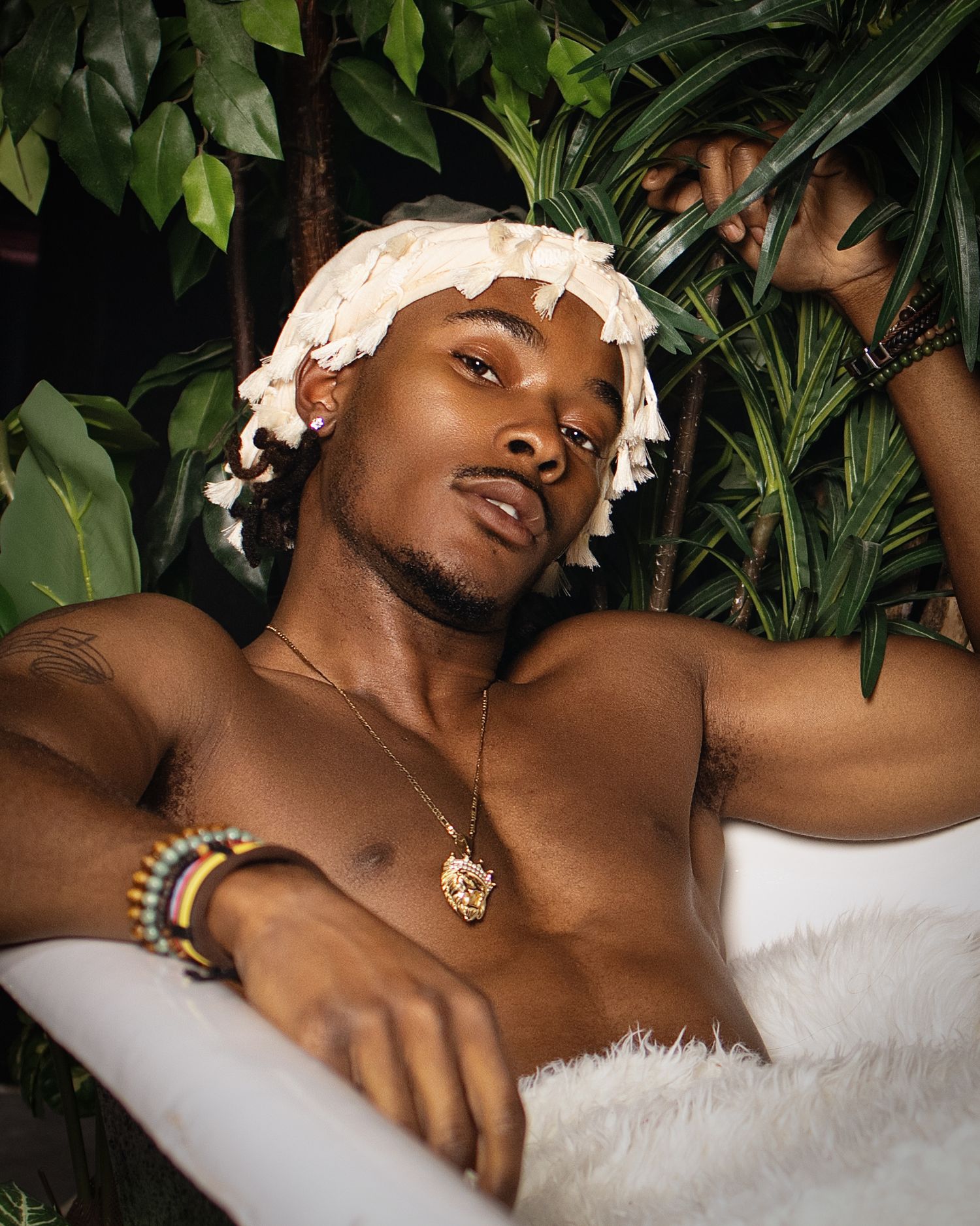Shirtless man relaxing in a bathtub surrounded by tropical plants, wearing gold jewelry and a white shower cap.