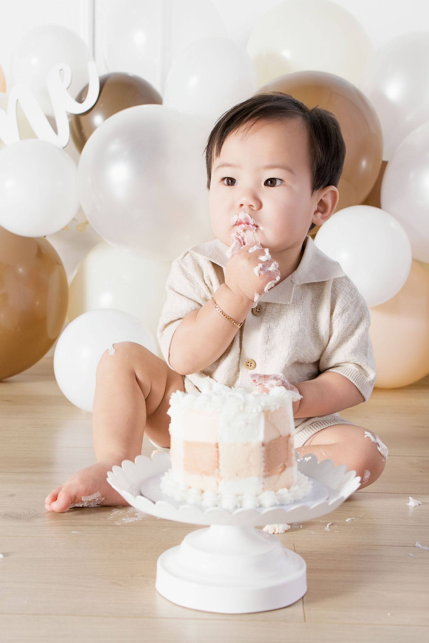 Baby boy in beige outfit eating first birthday cake surrounded by white and gold balloons on wooden floor.