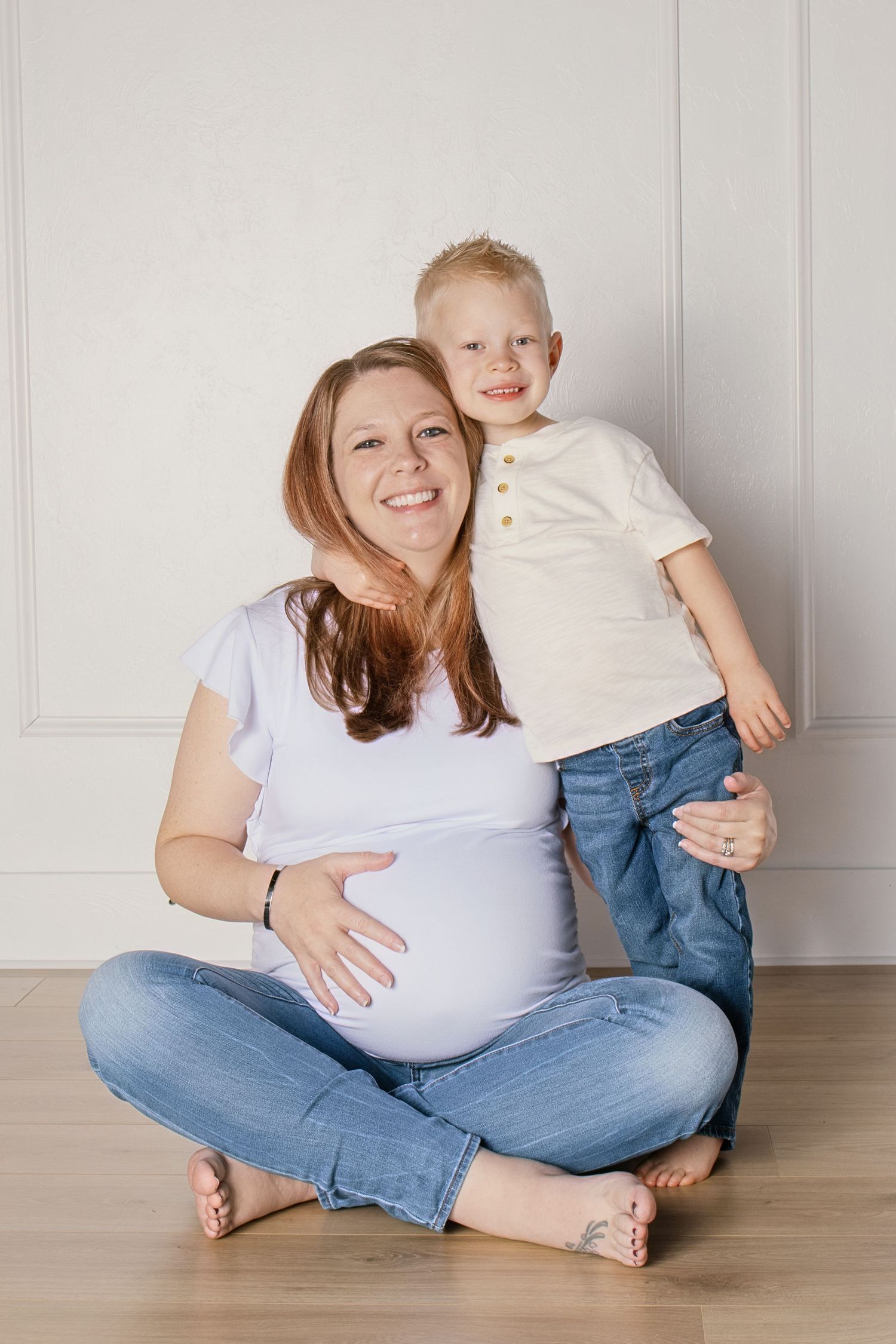 Pregnant woman sitting cross-legged on floor smiling with young blond boy hugging her from behind.