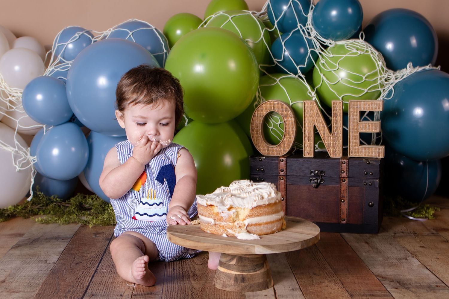 Baby's first birthday cake smash with blue and green balloons, ONE sign, and a white frosted cake on wooden floor.