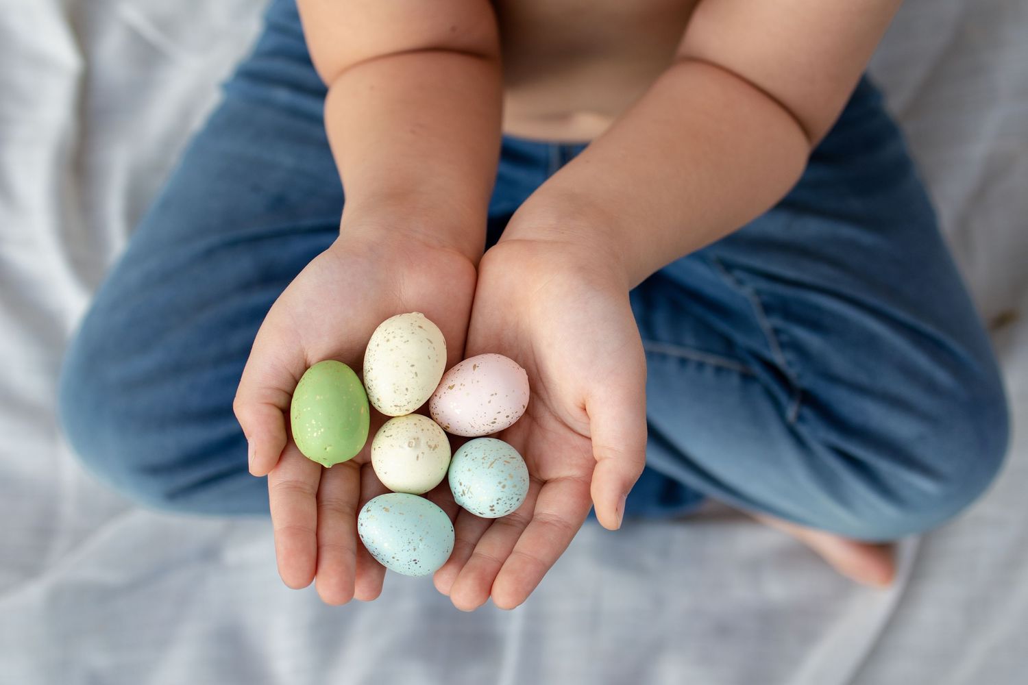 Child holding colorful pastel Easter eggs in small cupped hands, wearing blue jeans, sitting on white bedding.