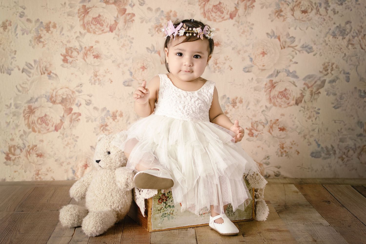 A baby girl in a white lace dress and floral headband sits on a vintage suitcase beside a teddy bear.