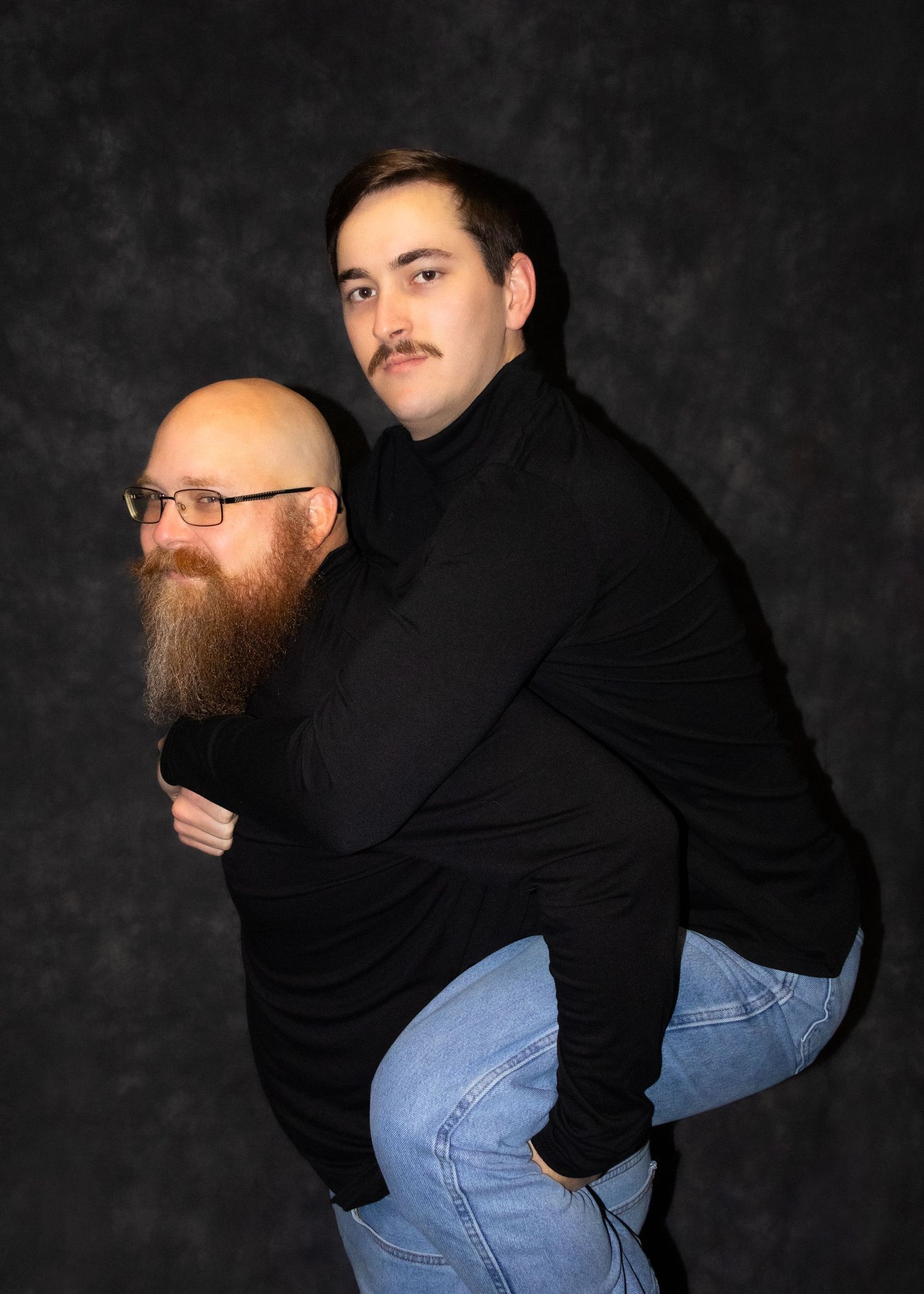 Two men wearing black turtlenecks pose in a studio, one giving the other a piggyback ride.