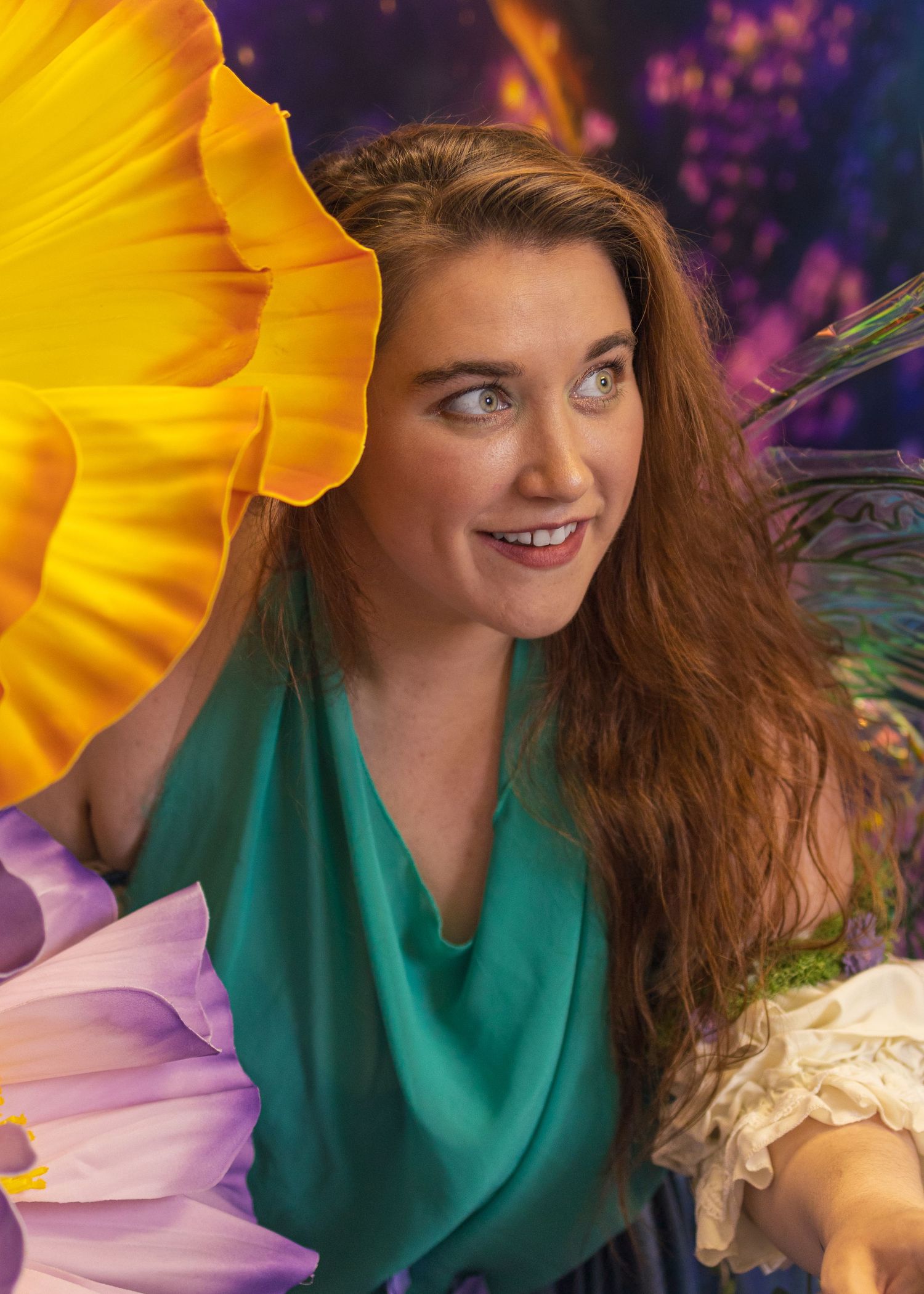 Woman in teal top smiling among oversized colorful flowers in a vibrant fantasy garden setting.