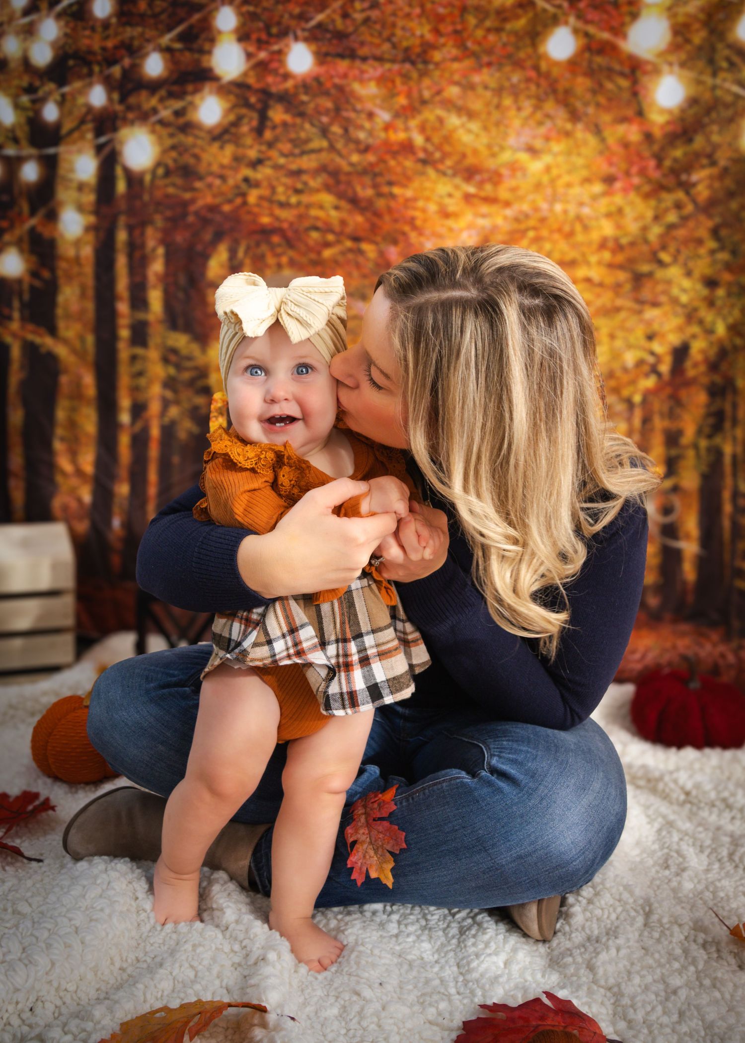 Mother kissing baby girl in fall autumn photo session with orange foliage backdrop and string lights.