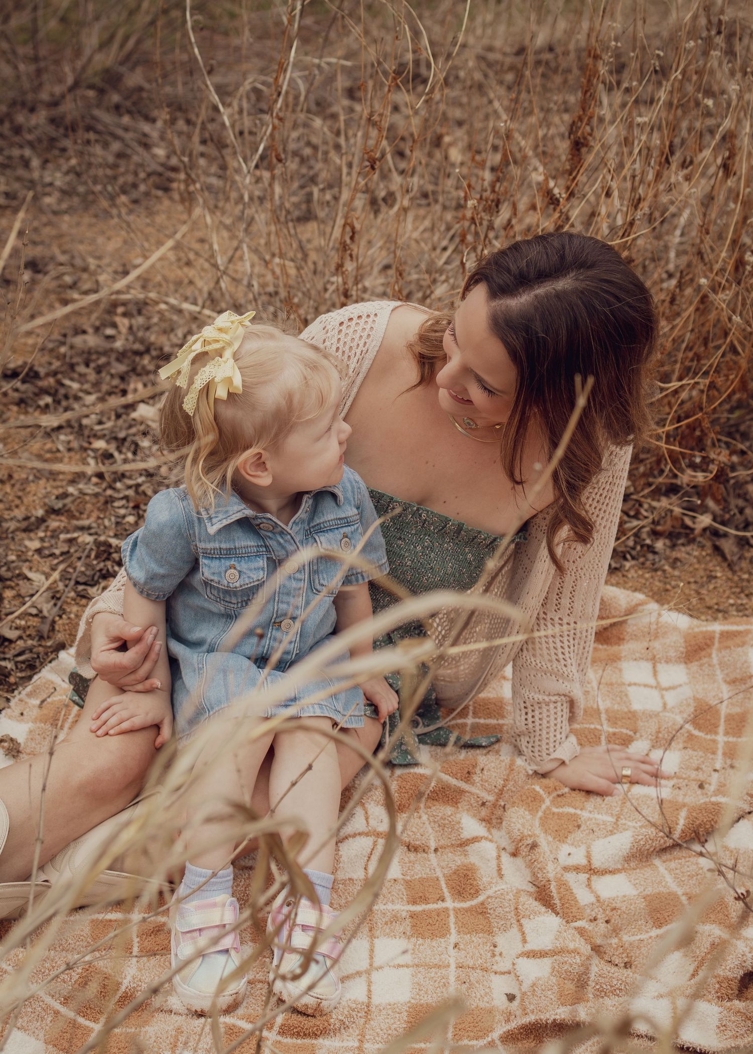 A mother and her young toddler daughter share a sweet nose-to-nose moment while sitting on a blanket outdoors.