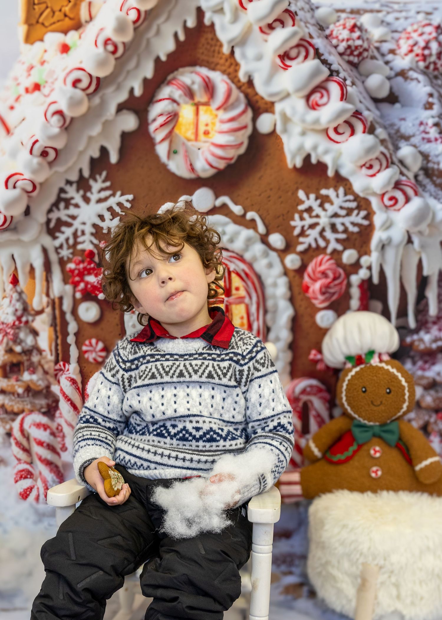 A curly-haired boy in a Nordic sweater sits before a festive gingerbread house with candy canes and icing decorations.