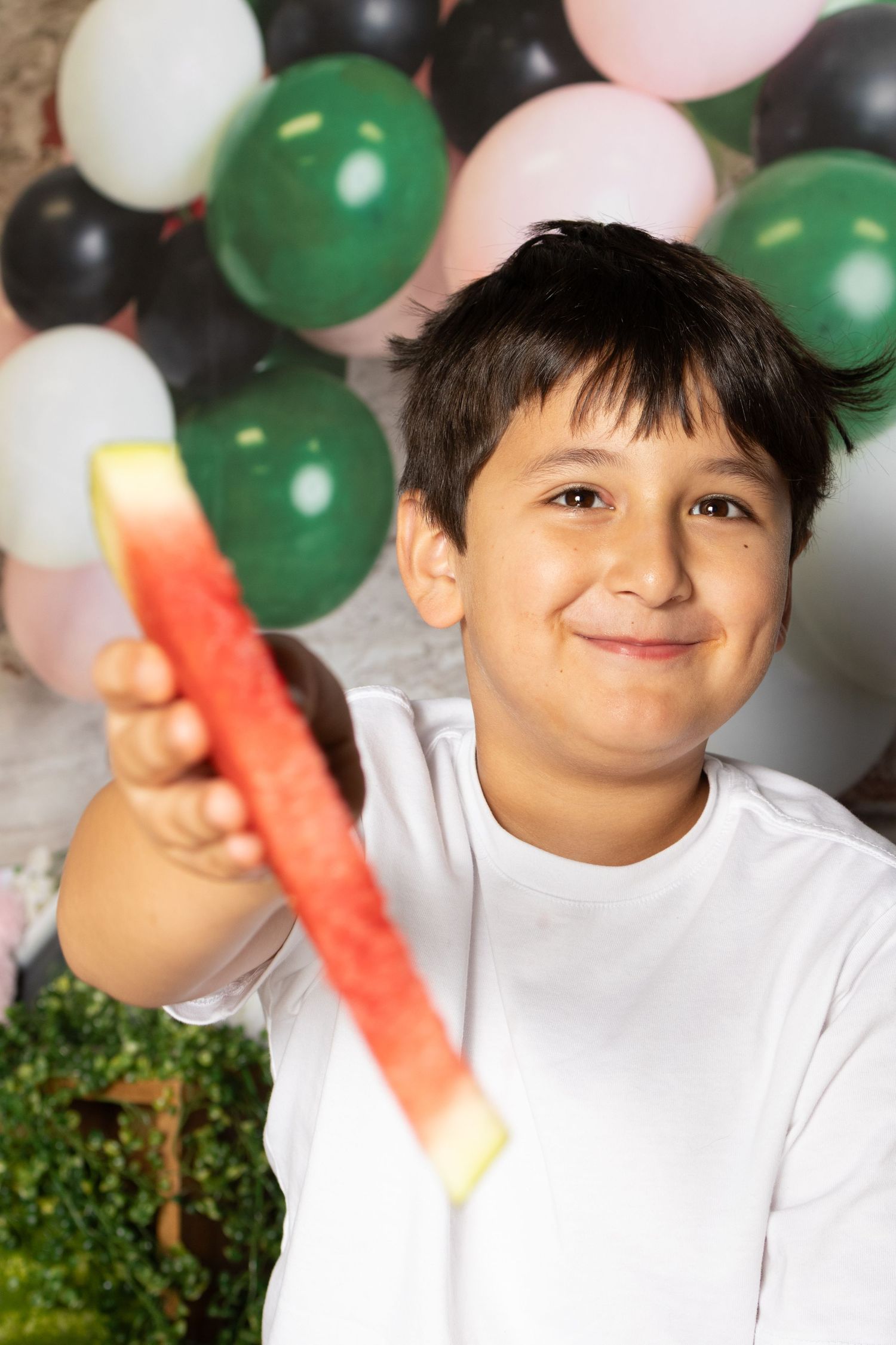 A smiling boy in a white shirt holds a watermelon slice toward the camera at a balloon-decorated party.