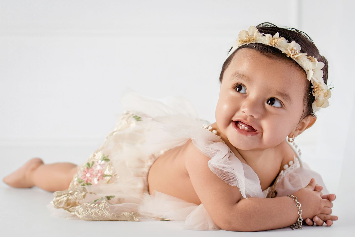 Adorable baby girl in a floral tutu dress and pearl bracelet with a flower crown, posing on a white background.