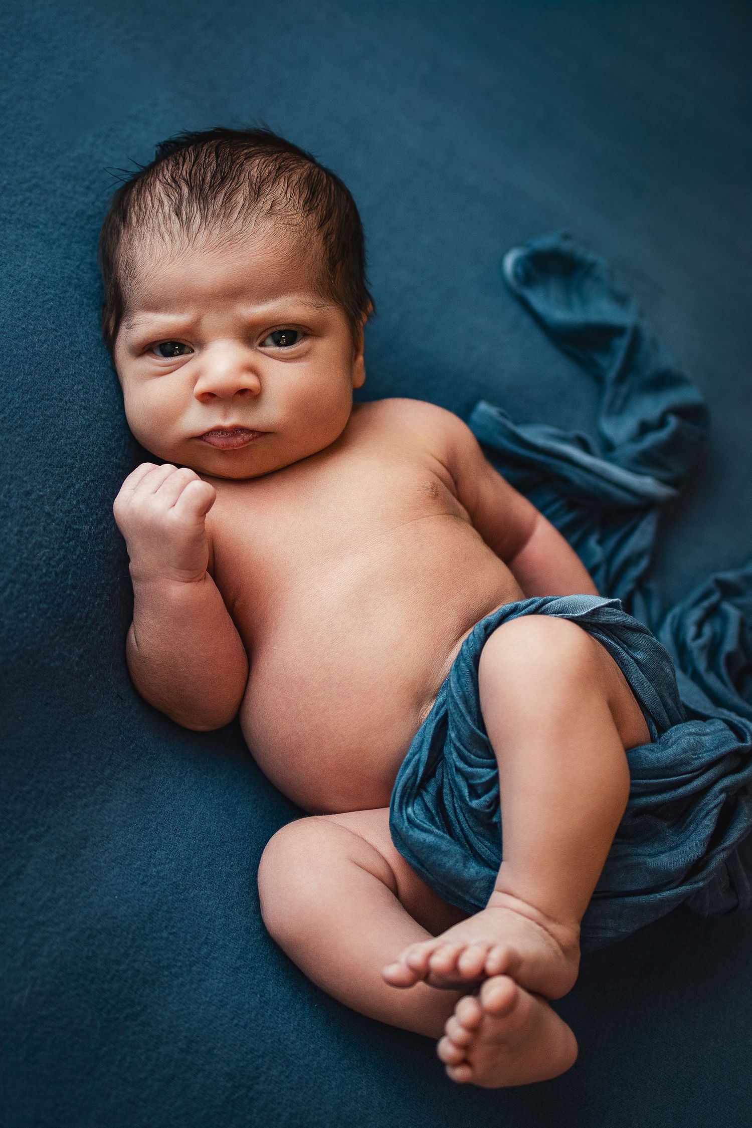 Newborn baby lying on a teal blanket, looking alert with a fist raised, wearing a blue wrap diaper.