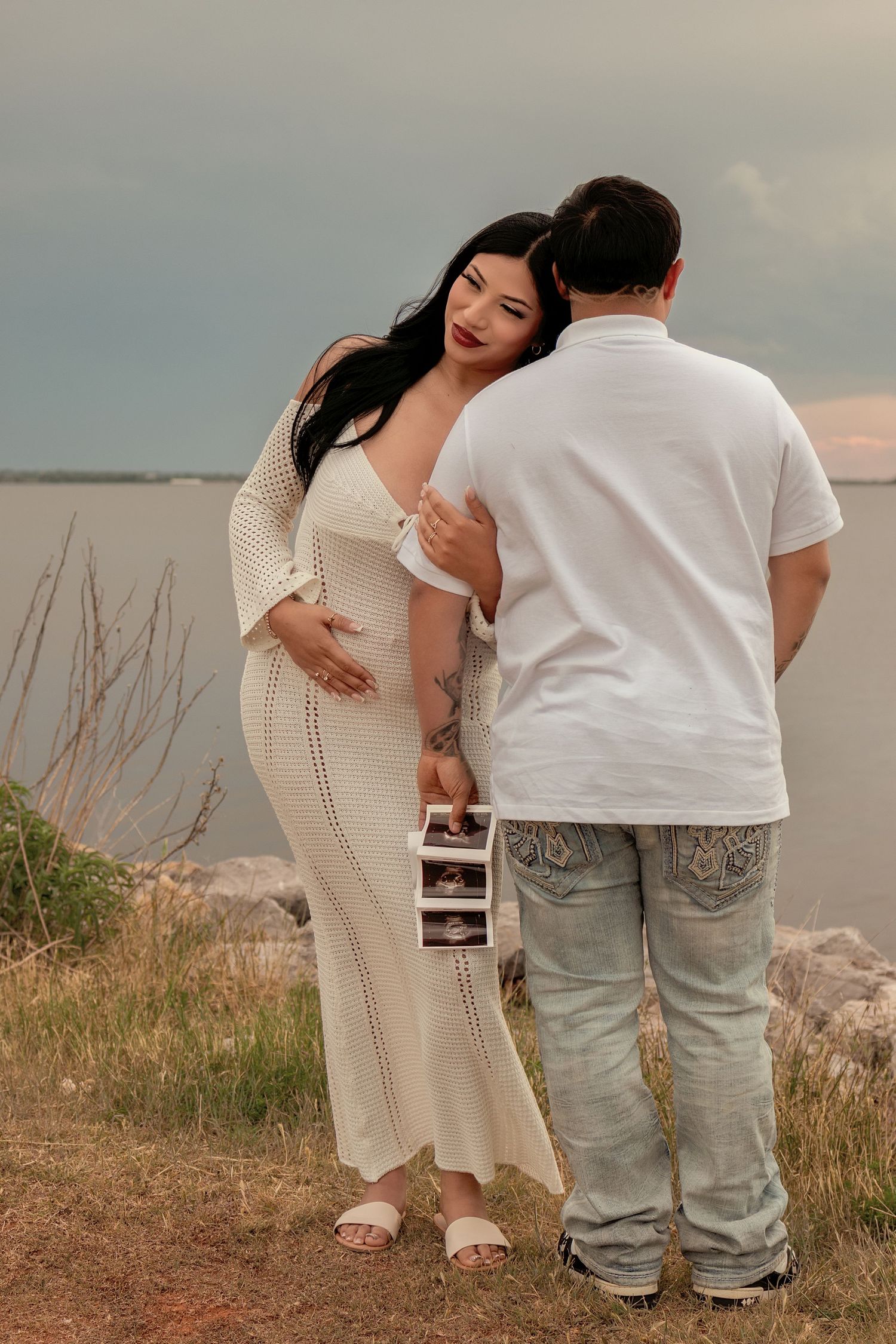 Pregnant woman in white dress holds ultrasound photos with partner by a lake during maternity photoshoot.