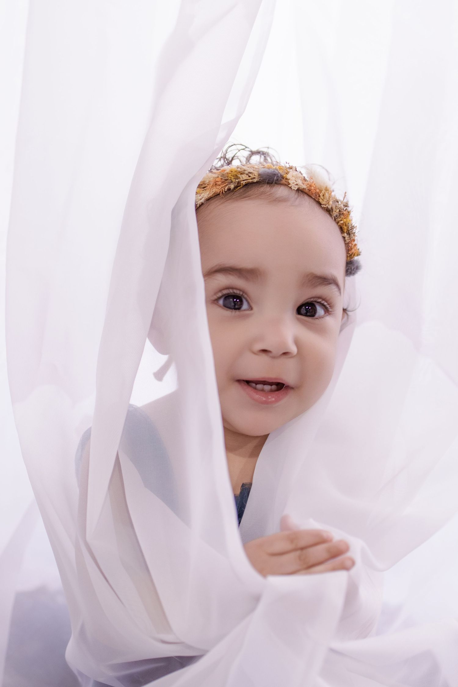 Adorable baby peeking through white sheer curtains wearing a golden crown headband.