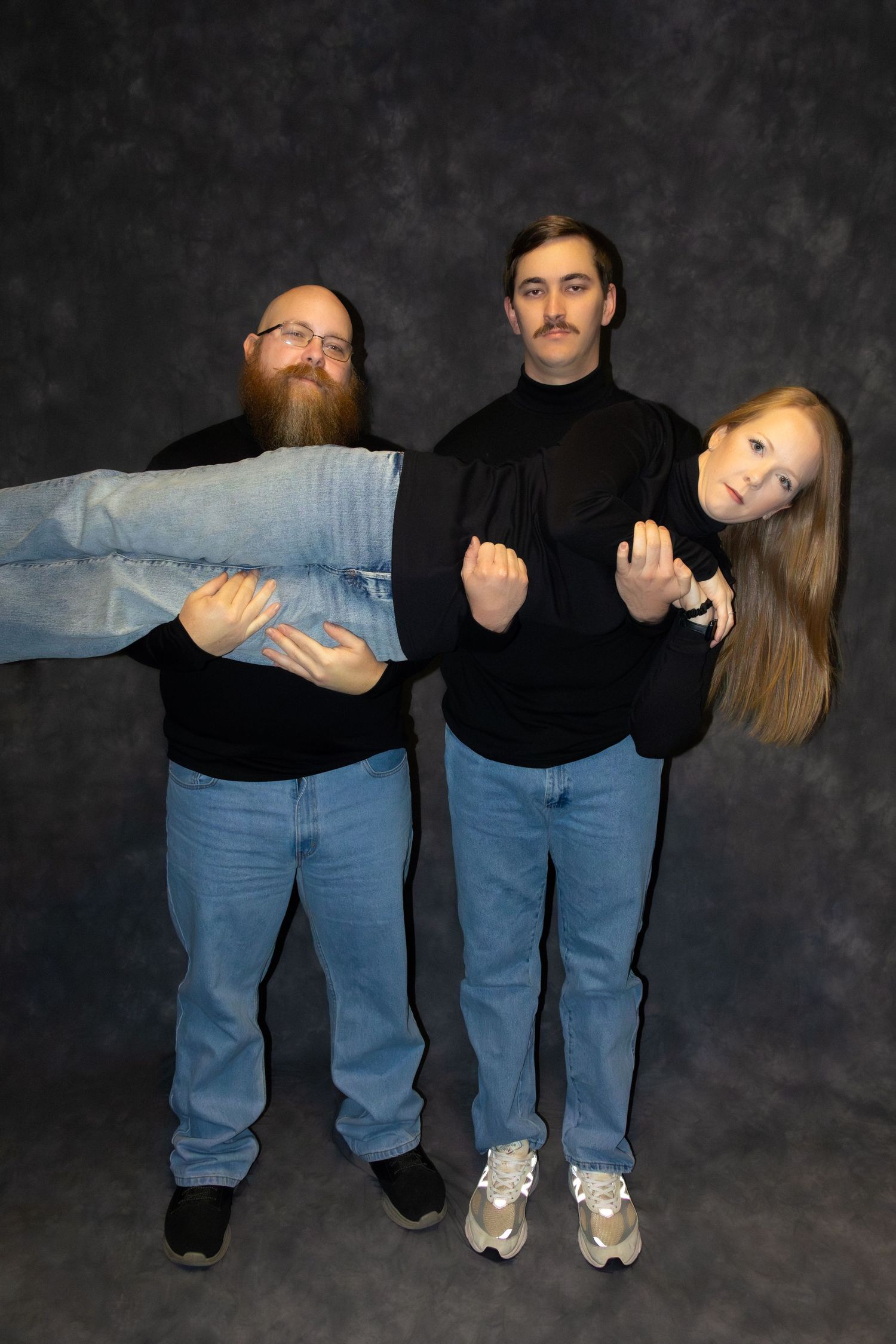 Three people in matching black turtlenecks and jeans pose creatively, with one person being held horizontally in a studio.