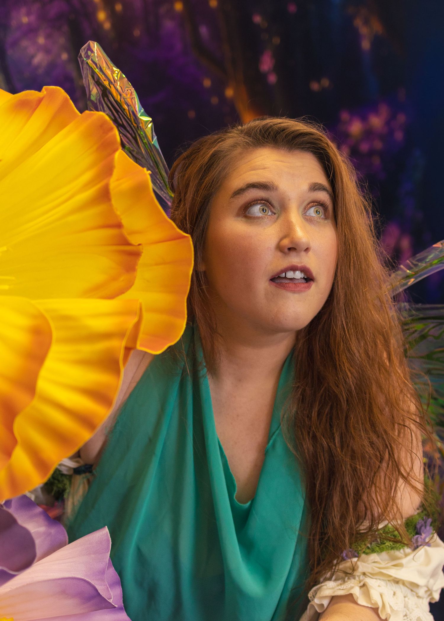 Woman in teal dress with fairy wings poses among colorful giant flowers in enchanted forest fantasy setting.