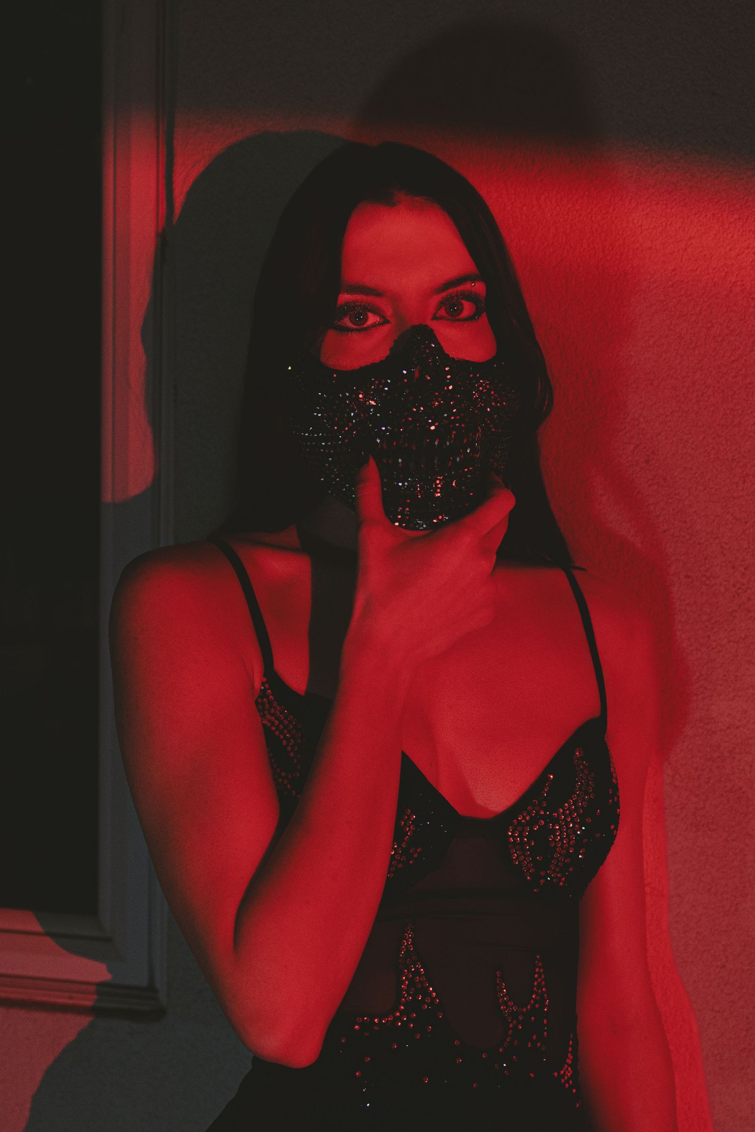Woman in black sequin mask and lingerie poses dramatically under red lighting in a dark, moody setting.