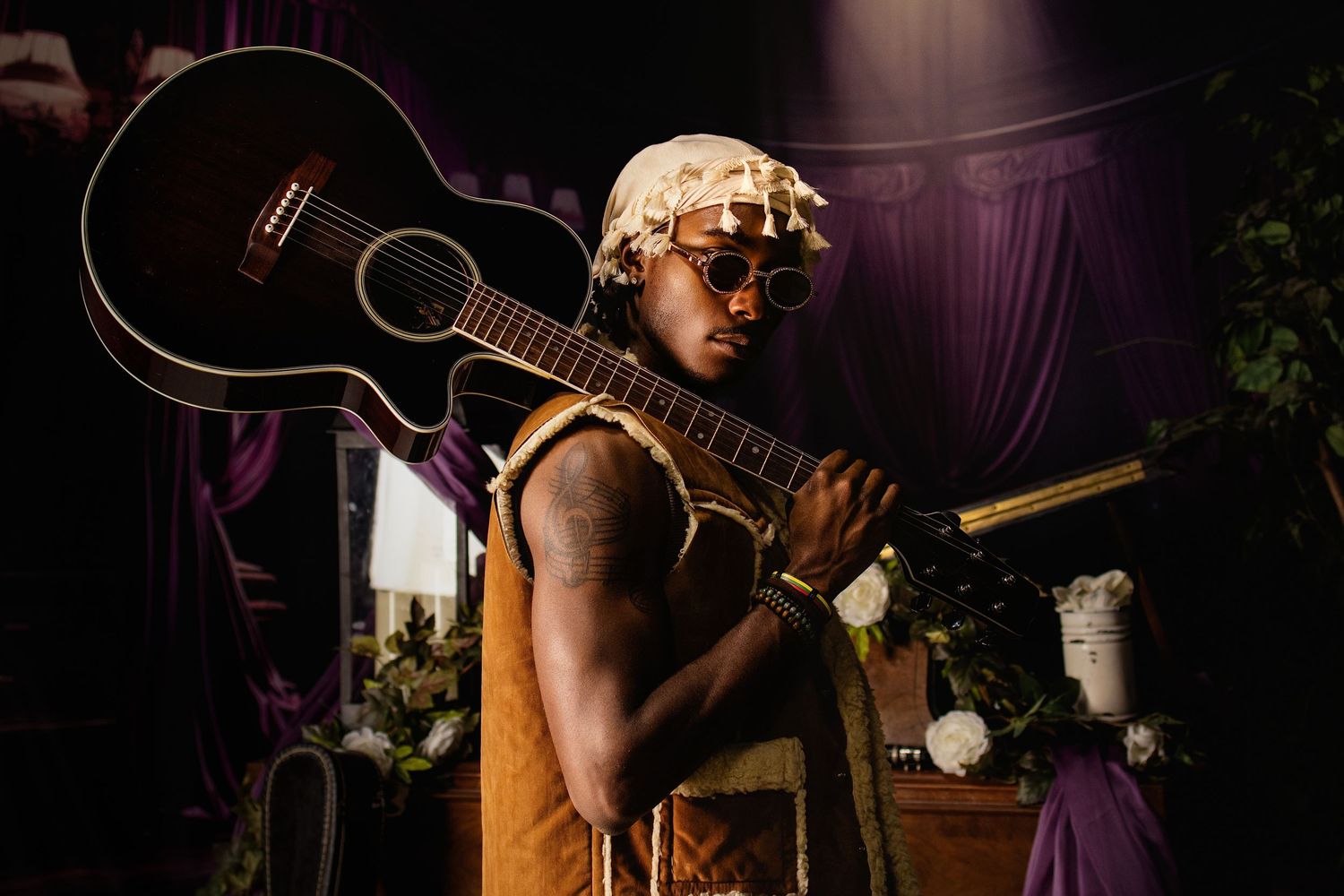 Musician holding acoustic guitar over shoulder wearing sunglasses and head wrap in dramatic stage lighting.
