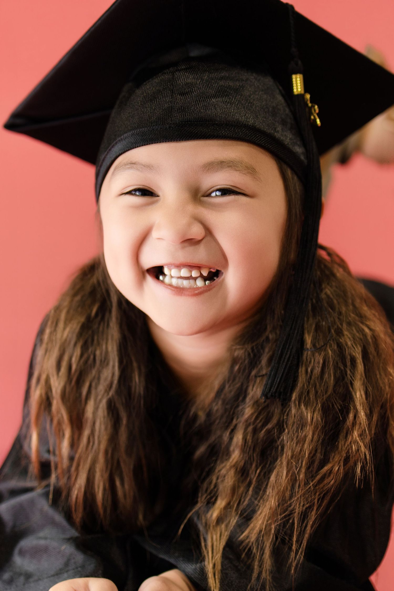 Happy young girl wearing black graduation cap and gown, smiling broadly against a pink background.