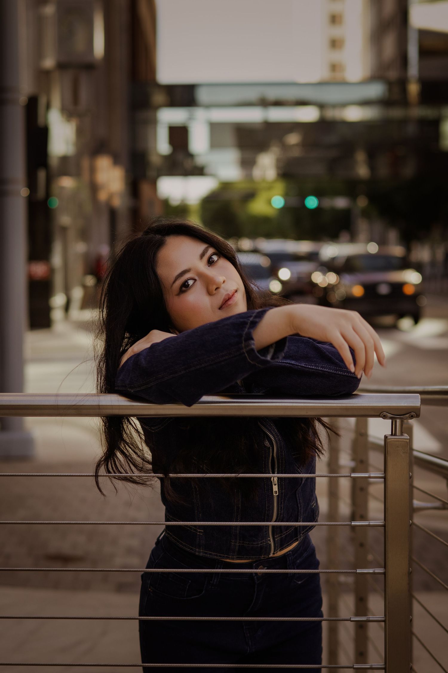 Young woman in navy velvet jacket leans thoughtfully on metal railing on urban city street at dusk.