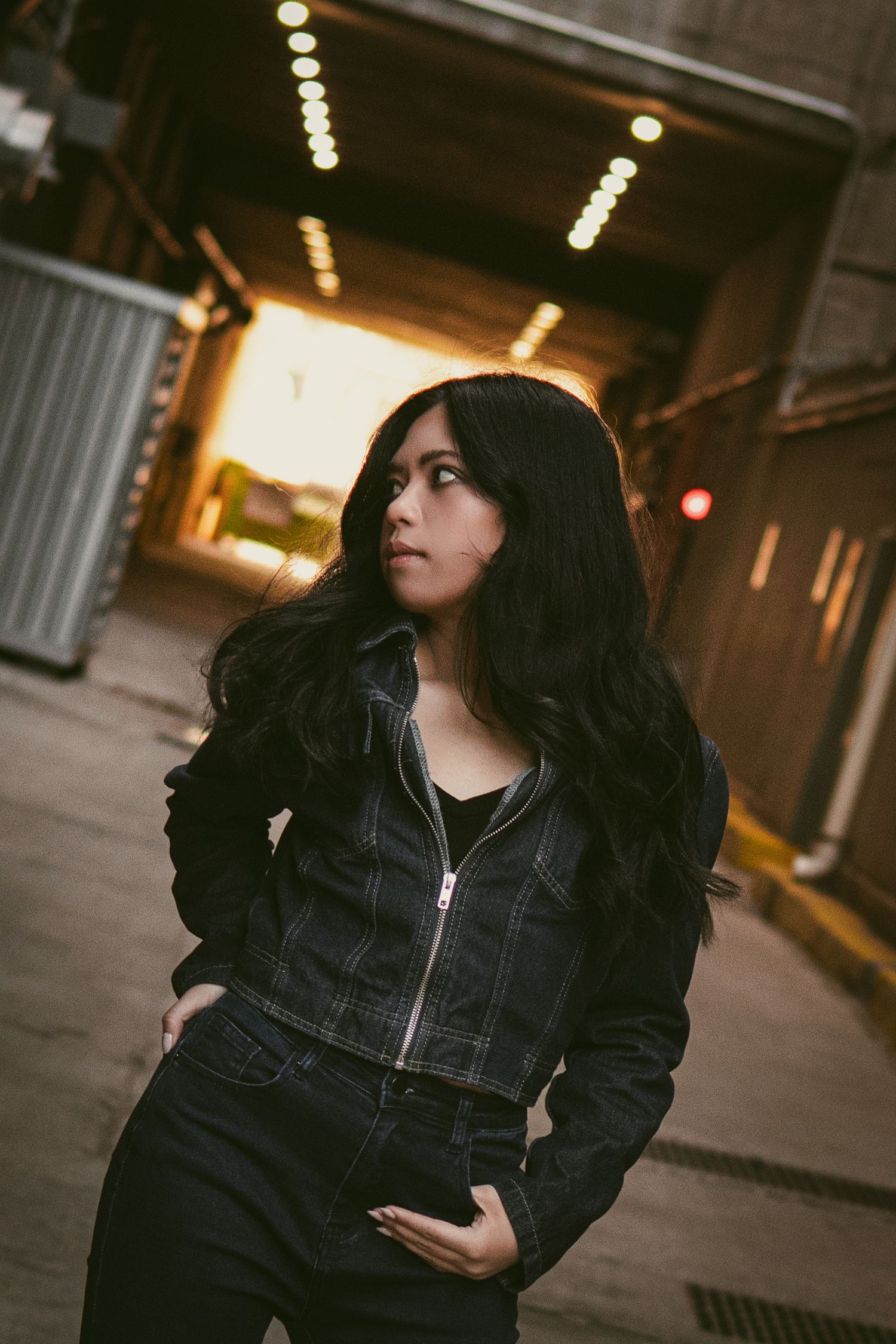 Young woman with long black hair in dark denim jacket stands in urban alley with glowing tunnel behind her.