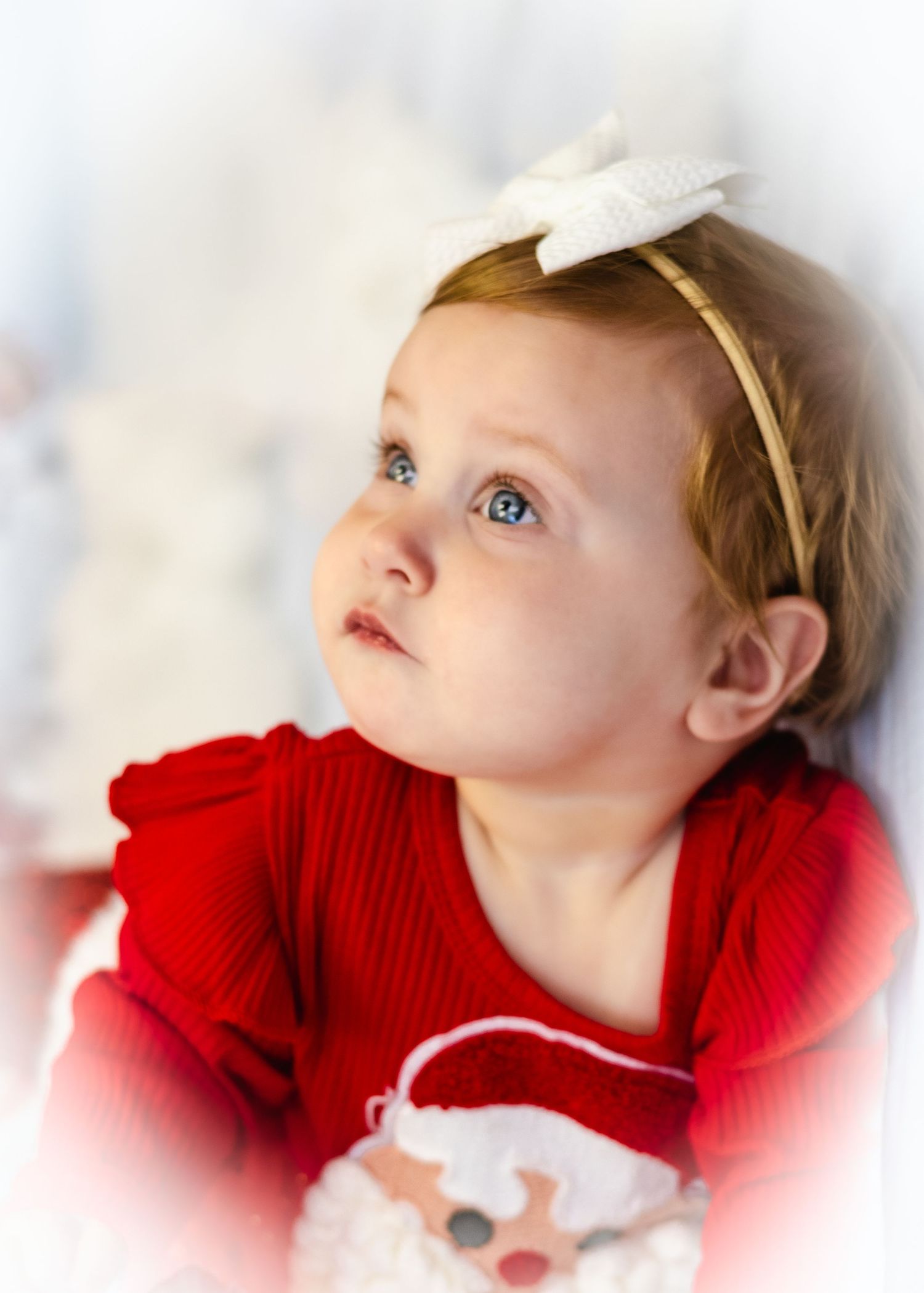 A toddler girl in a red Christmas outfit and white bow headband gazes upward with bright blue eyes.