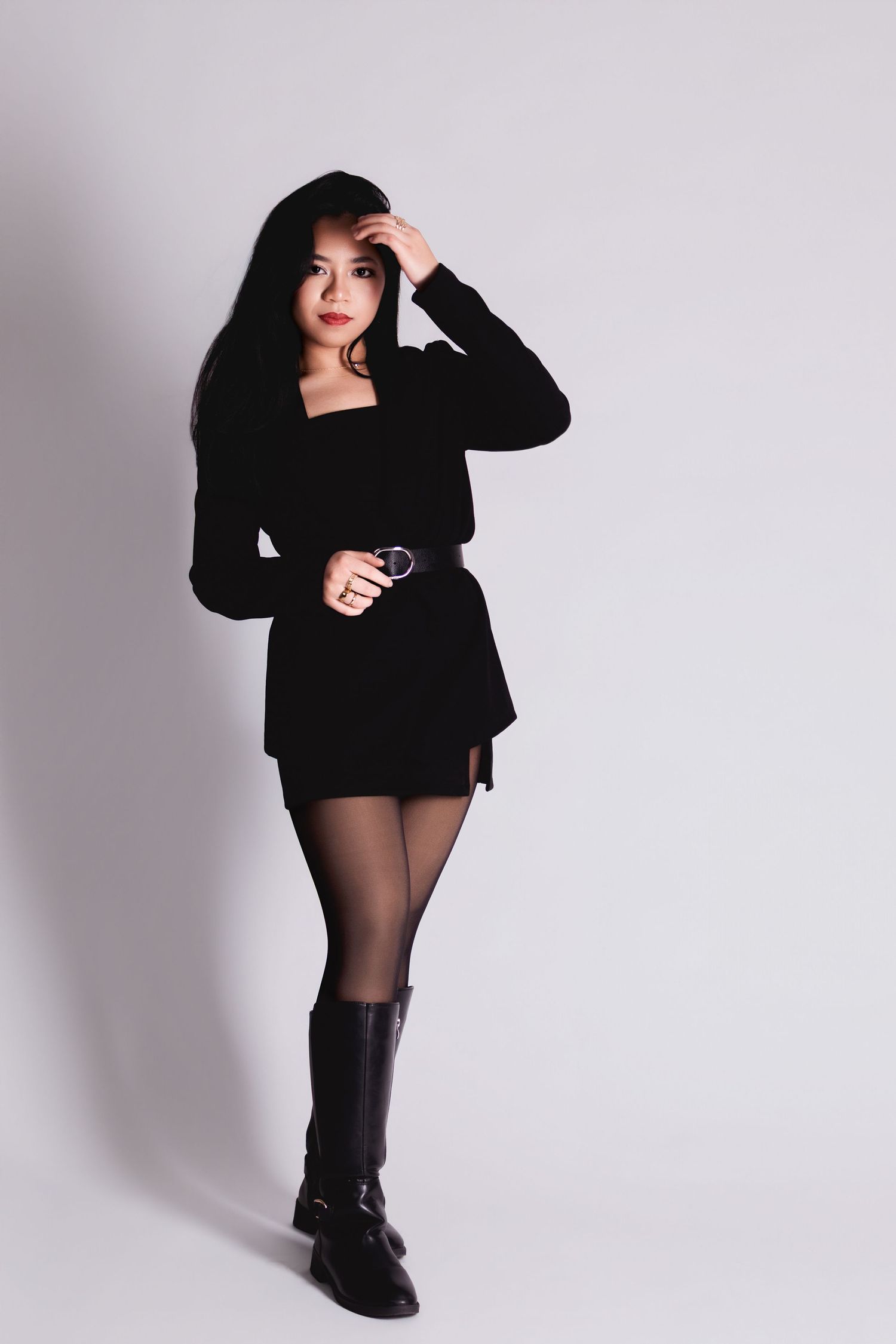 Woman in black mini dress, belt, tights, and knee-high boots poses confidently against a light gray background.