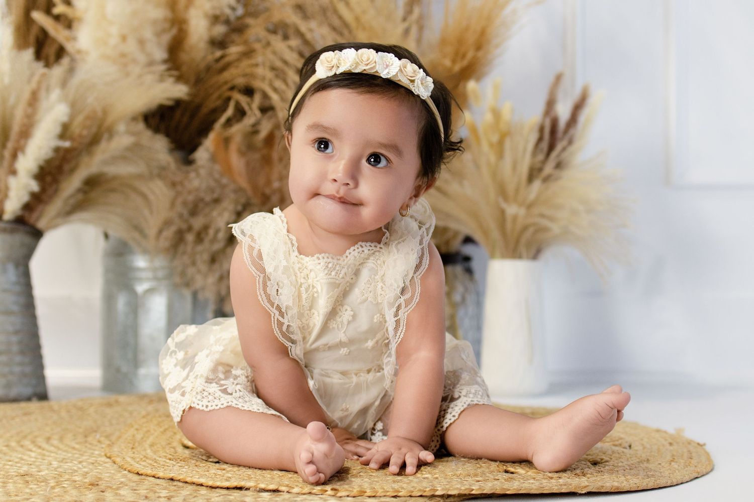 Baby girl in cream lace romper and floral headband sits on woven rug with pampas grass decor.