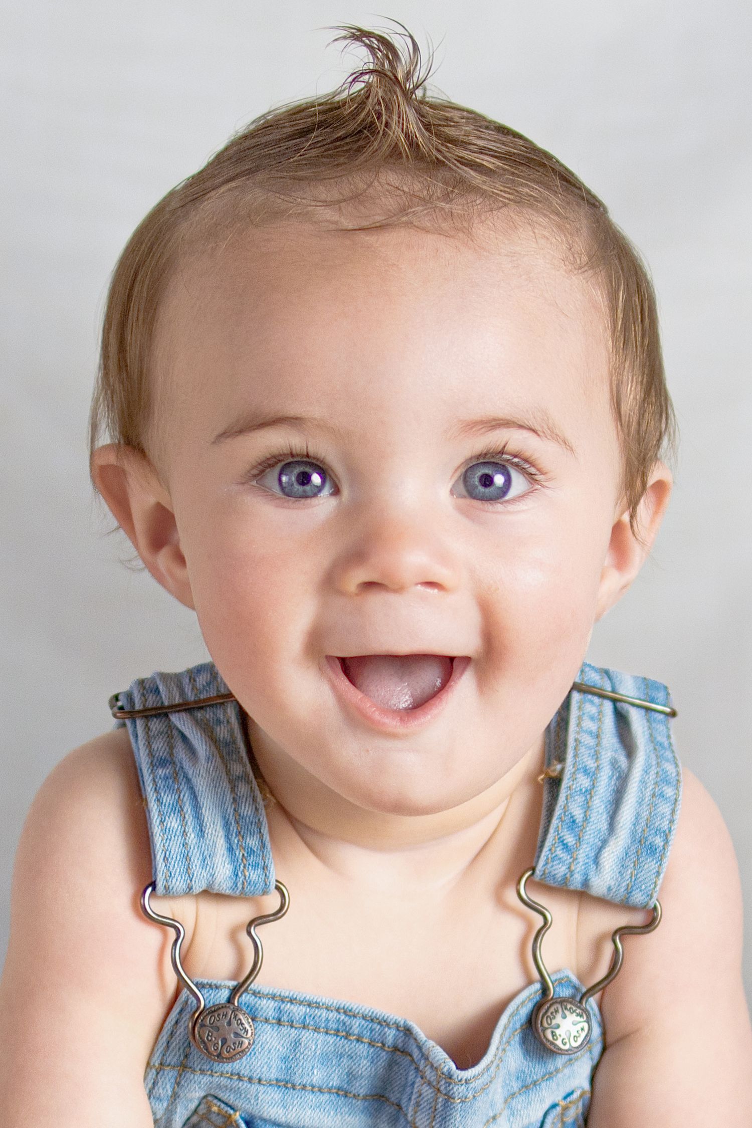 Adorable smiling baby with blue eyes wearing denim overalls, portrait photo on gray background.