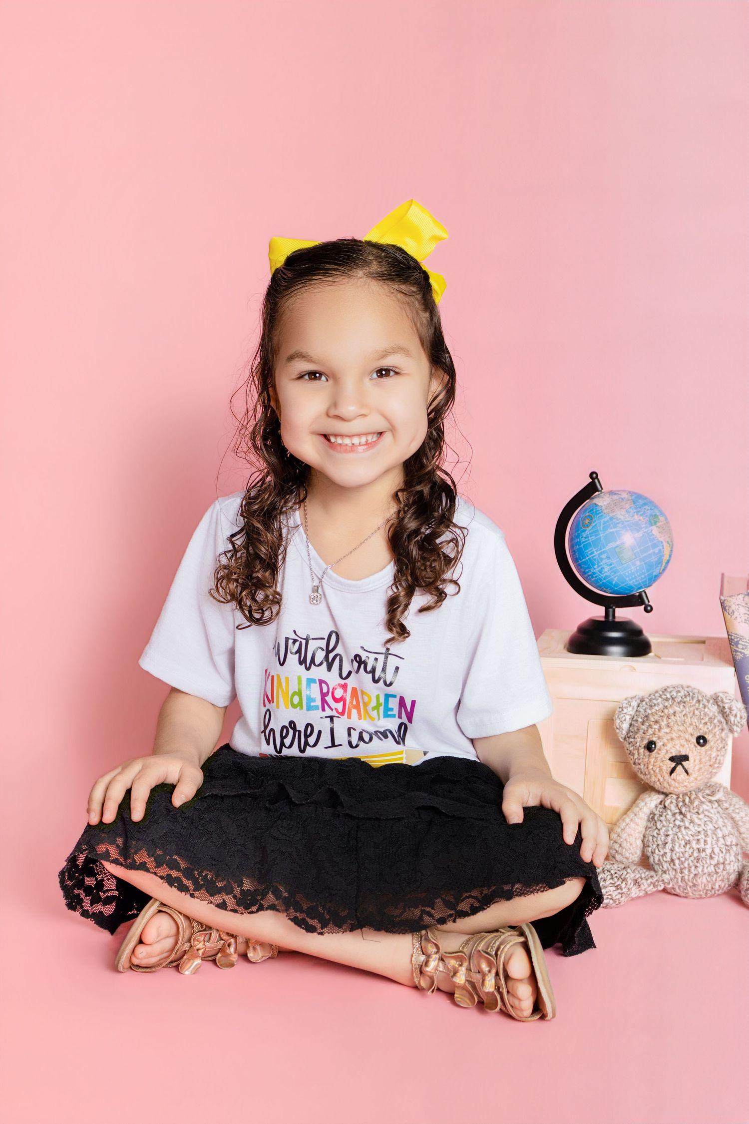 Smiling girl in kindergarten shirt sits cross-legged with globe and teddy bear on pink background.