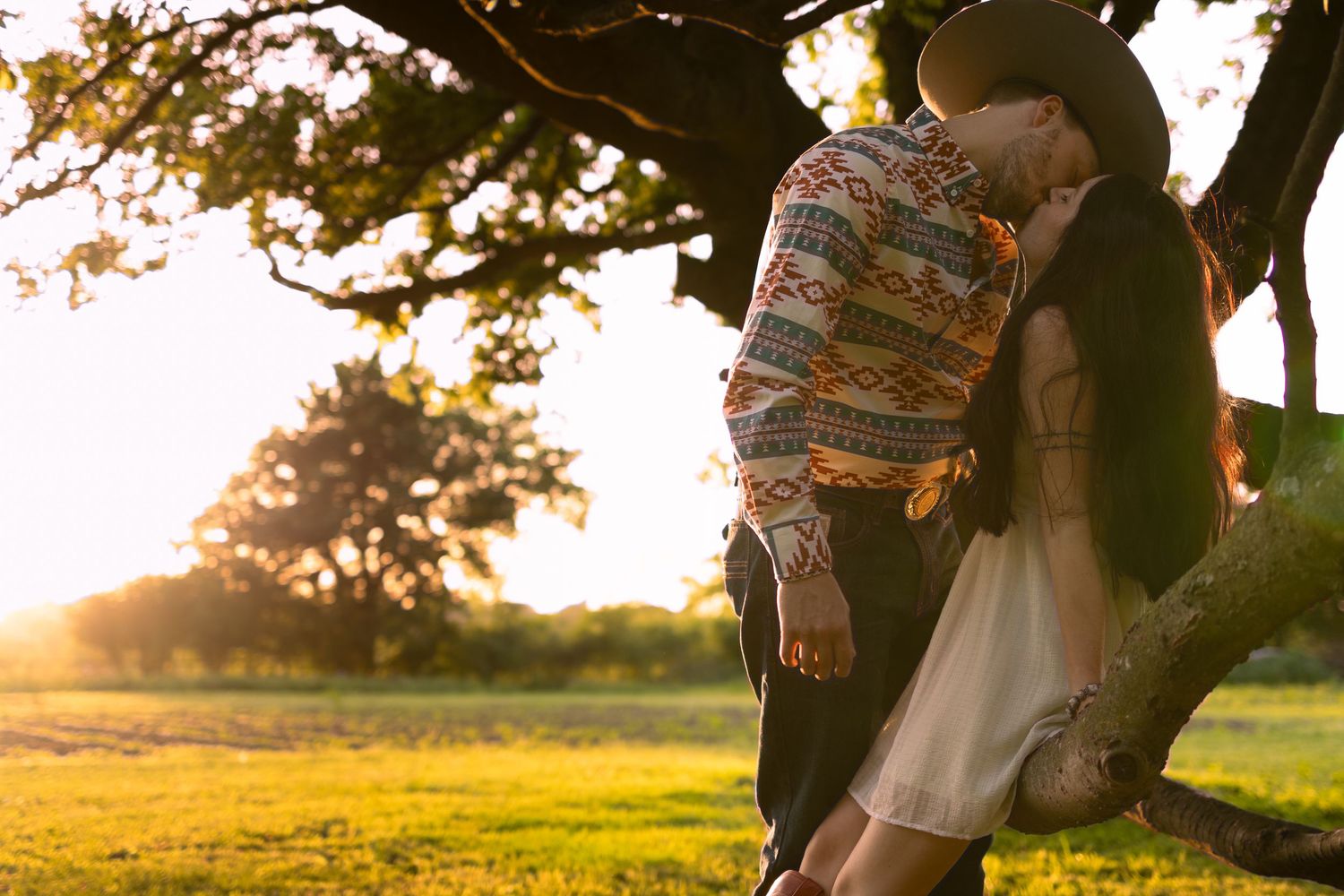 Couple sharing a romantic kiss under a tree at golden sunset in a lush green park.