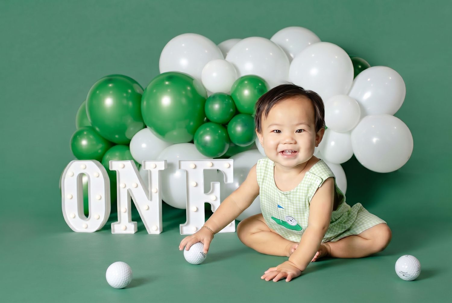 Smiling baby sits before green and white balloon garland with ONE marquee letters on a green backdrop.