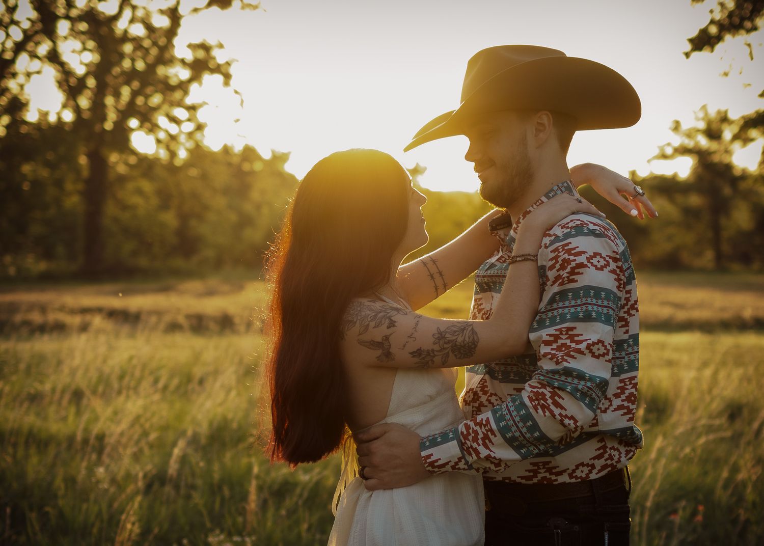 Couple embracing at golden hour in a field, man wearing cowboy hat and aztec shirt, woman with tattoos in white dress.