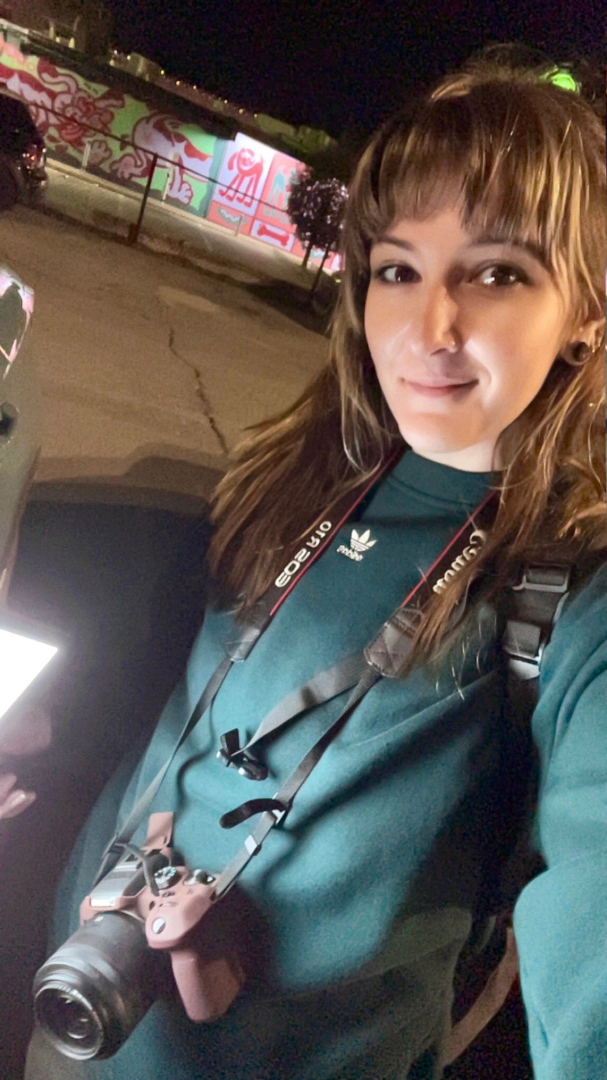 Woman in teal Adidas sweatshirt holding a Canon camera with strap, taking a selfie outdoors at night near graffiti wall.